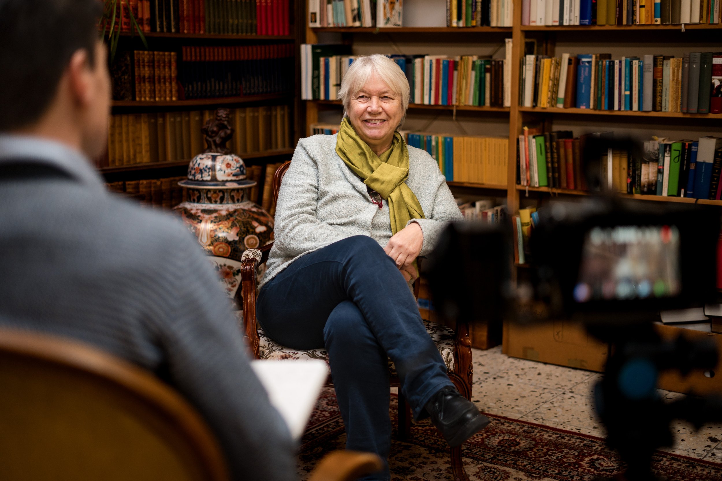 Eine ältere Frau sitzt in einem Bücherregalraum und spricht in einem Interview, während eine Person mit einem Notizblock und ein Kameraaufnehmen sie beobachten.