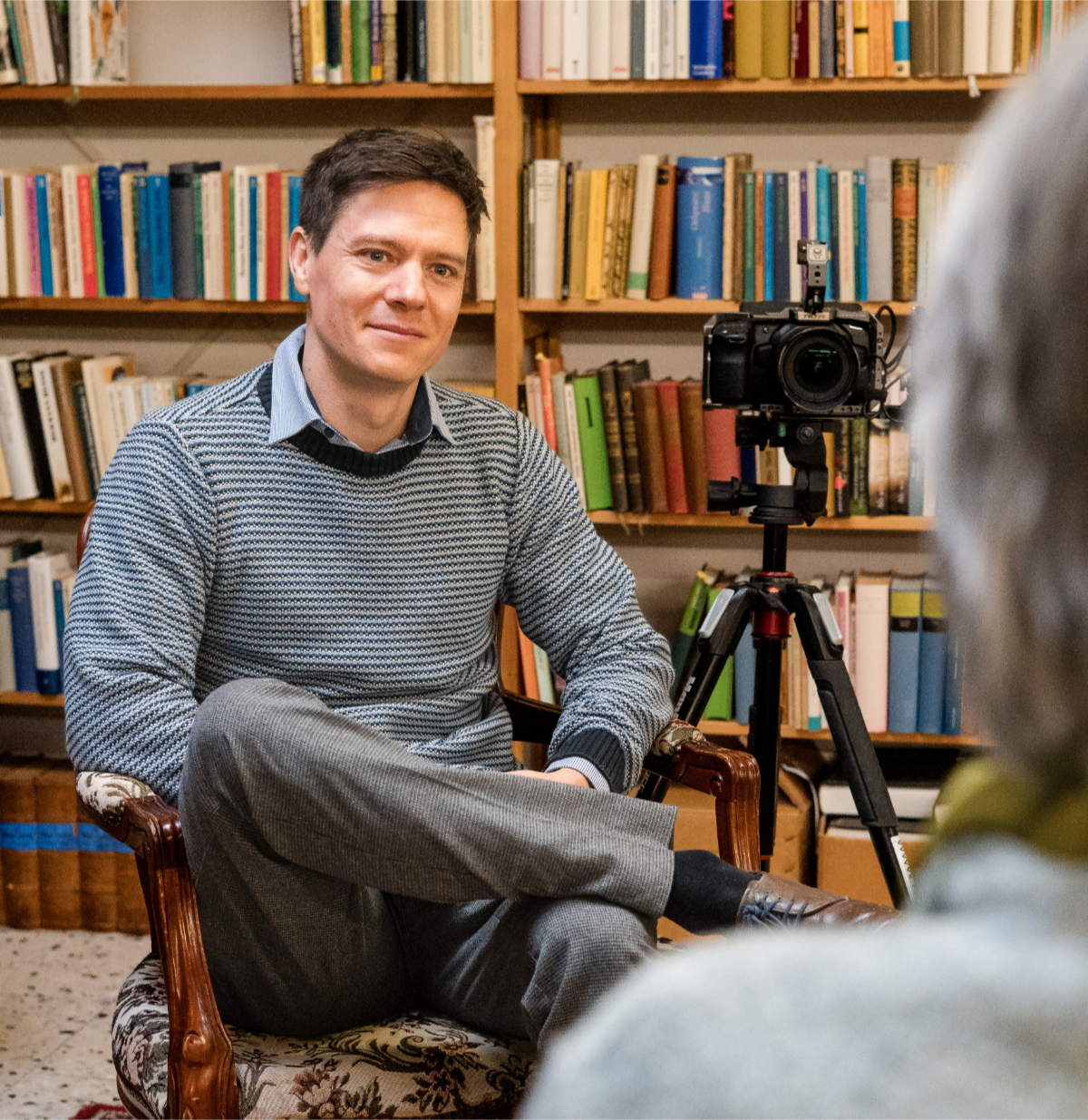 Ein Mann sitzt in einem Raum vor einer Kamera, im Hintergrund sind Bücherregale mit bunten Büchern.