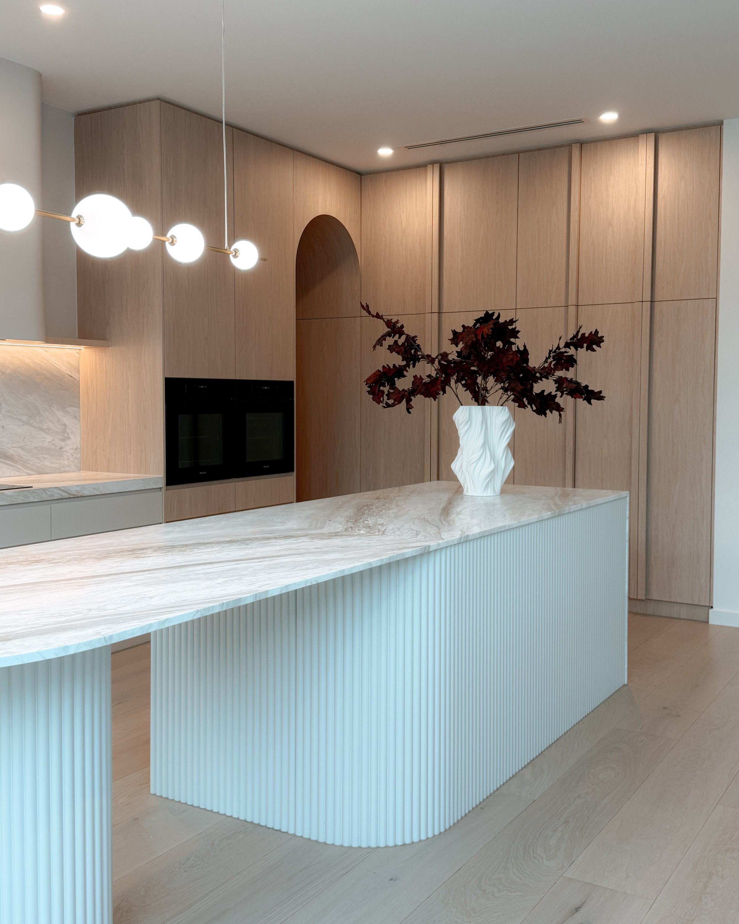 Contemporary soft-curved kitchen with fluted island, light timber cabinetry, and sculptural rangehood in a minimal-luxe Melbourne new-build. Designed by TID Studio, interior design in Sydney