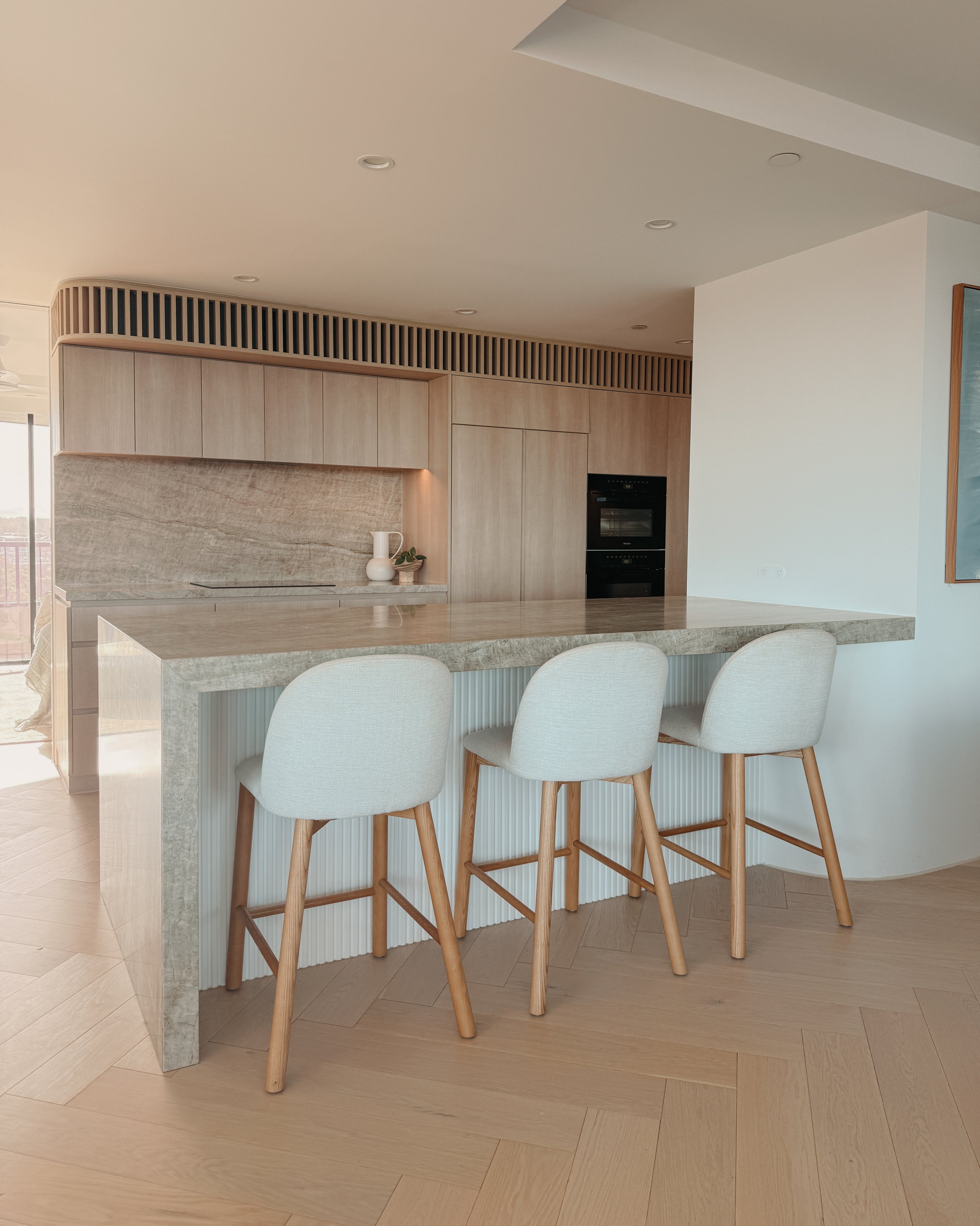 Contemporary condo renovation, modern coastal kitchen with curved island in Taj Mahal stone and pale timber cabinetry in Oceanview Apartment in Gold Coast. Designed by TID Studio Sydney