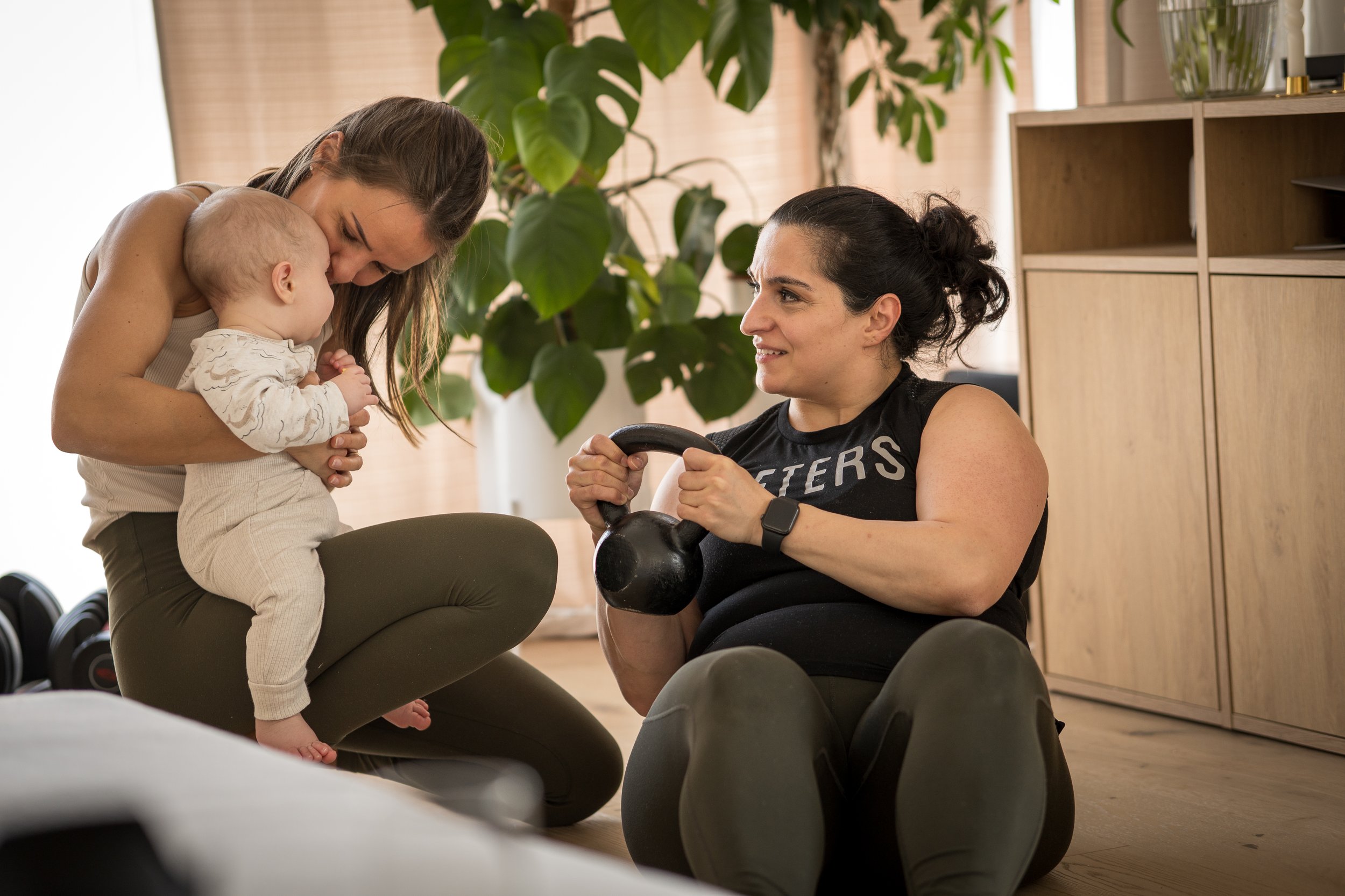 Eine Mutter trainiert ihre Bauchmuskulatur nach Geburt. Ihr Kind sitzt auf dem Schoßß der Trainerin und ist zufrieden, so wie die Mutter.