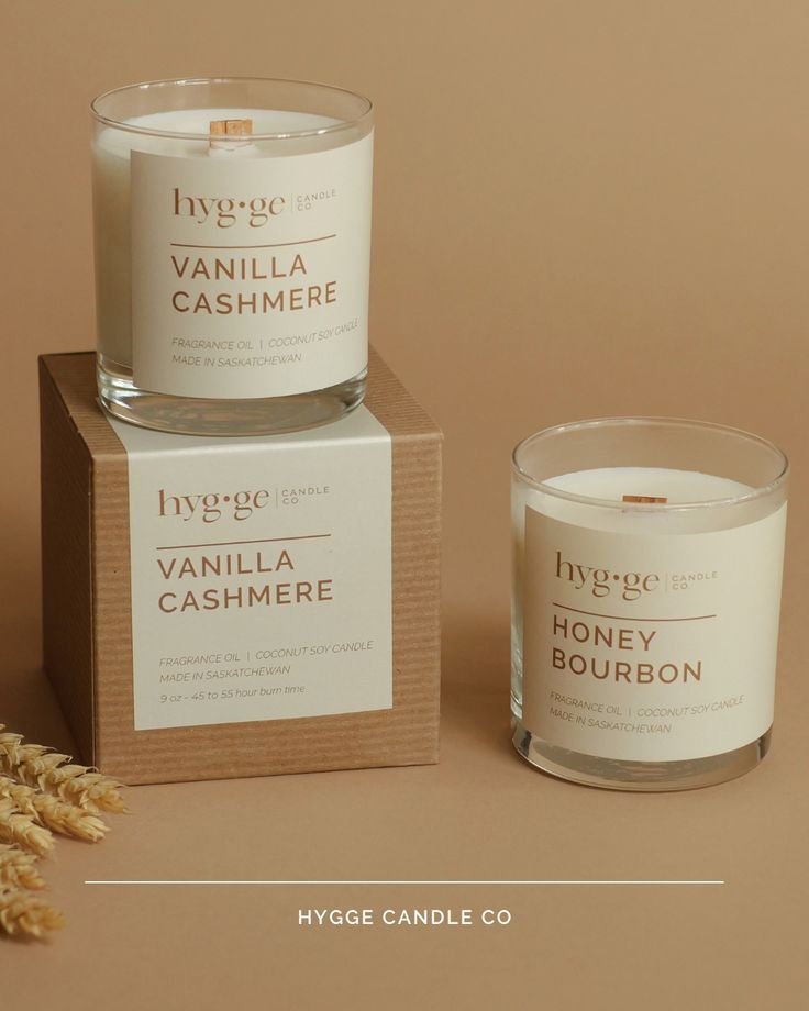 Two white scented candles from Hygge Candle Co, one labeled Vanilla Cashmere and the other Honey Bourbon, with the Vanilla Cashmere candle placed on a cardboard box and the Honey Bourbon candle beside it.
