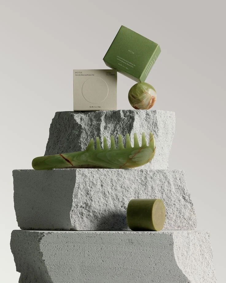 A minimalist display of green marble objects, including a spherical object, a comb-shaped object, and a cylindrical object, arranged on top of large textured gray concrete blocks with a small green box and a white box in the background.