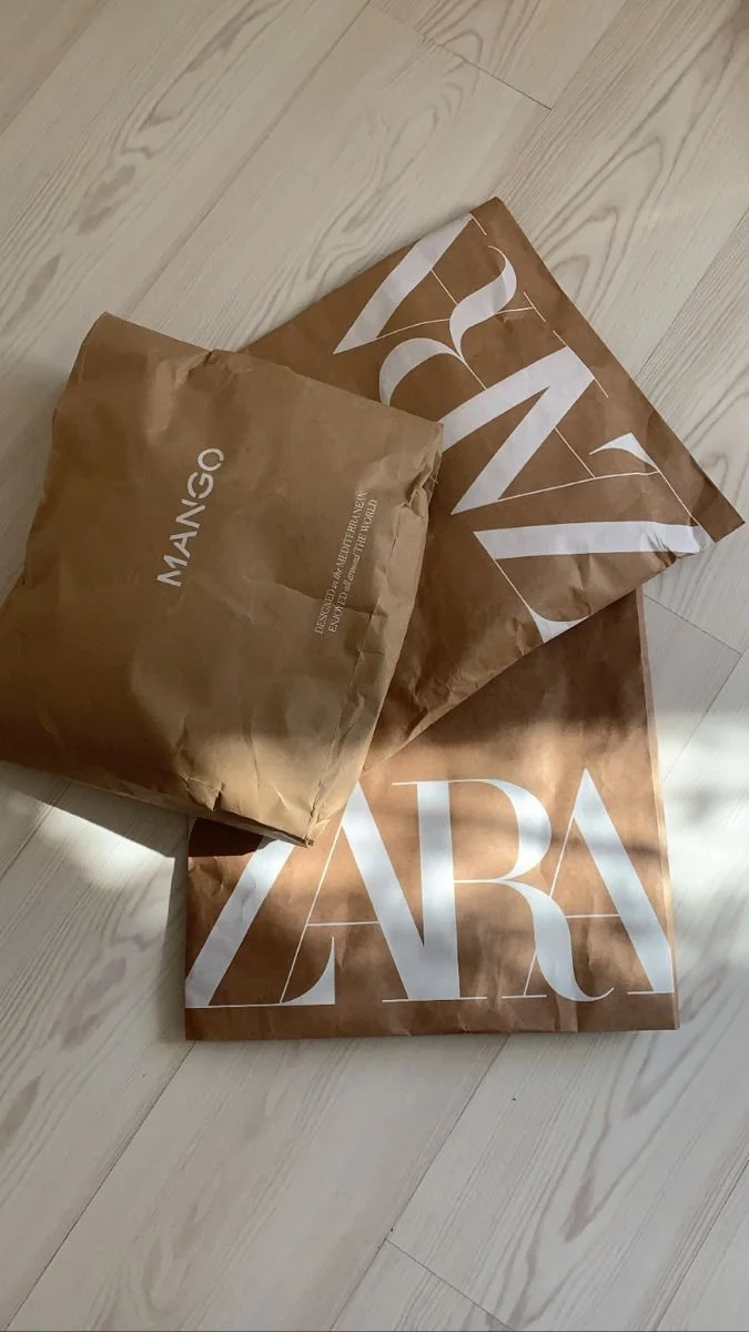 Three Zara shopping bags on a wooden floor, two large and one smaller, all made of brown paper with white Zara branding.