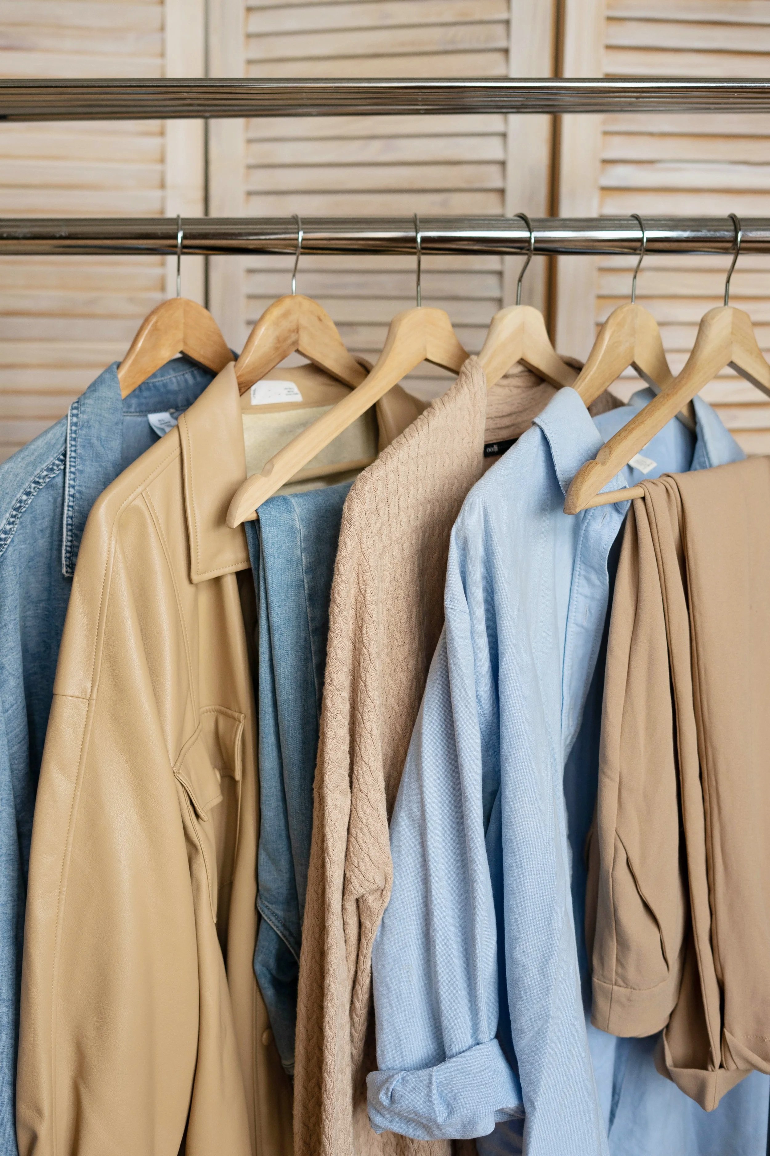 Clothing rack with various clothes including denim, beige jacket, beige sweater, light blue shirt, beige pants on wooden hangers in front of wooden slatted background.