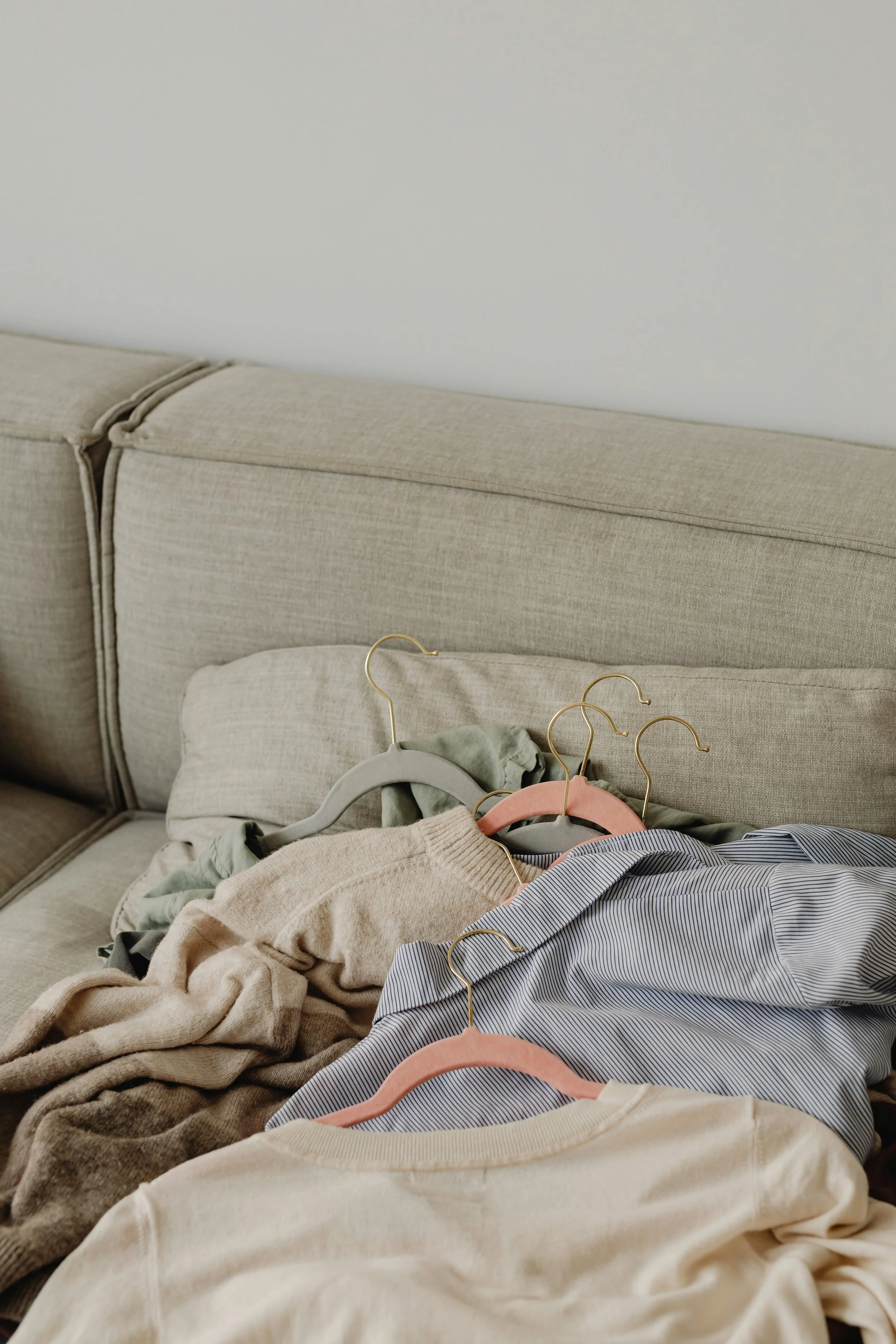 A beige sofa with a pile of clothes on it. The clothes include a beige sweater, a blue and white striped shirt, a green shirt, and a cream-colored shirt. The clothes are on hanging hangers, some with pink and gray hooks.