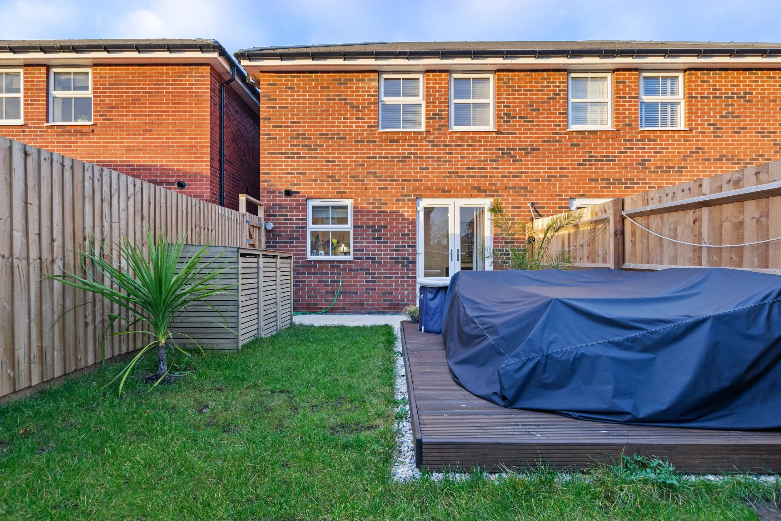 Backyard with grass lawn, wooden deck, covered hot tub, potted plants, and fence, with a brick house in the background.