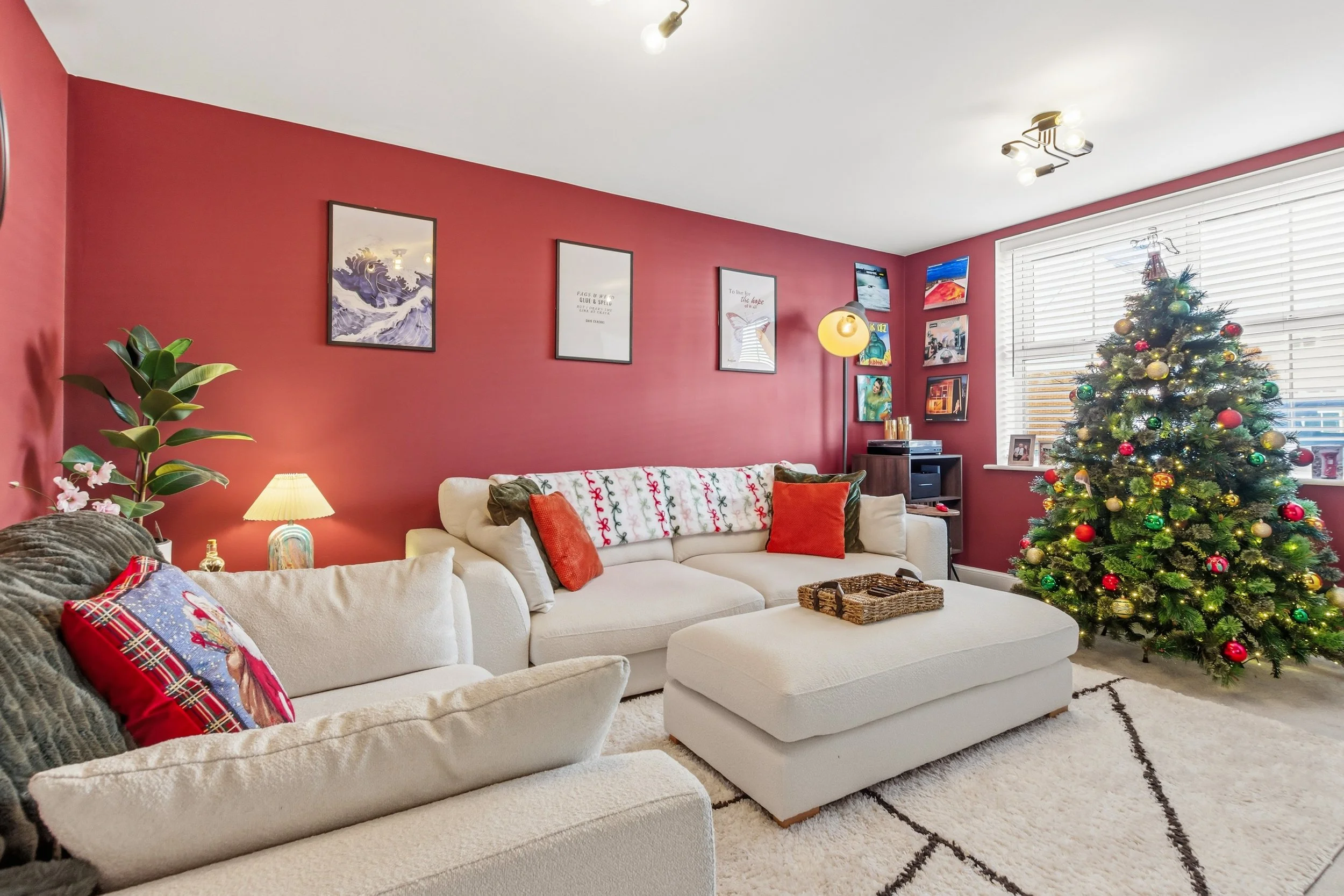 Living room decorated for Christmas with a decorated Christmas tree, white couches with red and green pillows, a white rug, a pink accent wall with framed artwork, a window with blinds, and festive holiday decor.