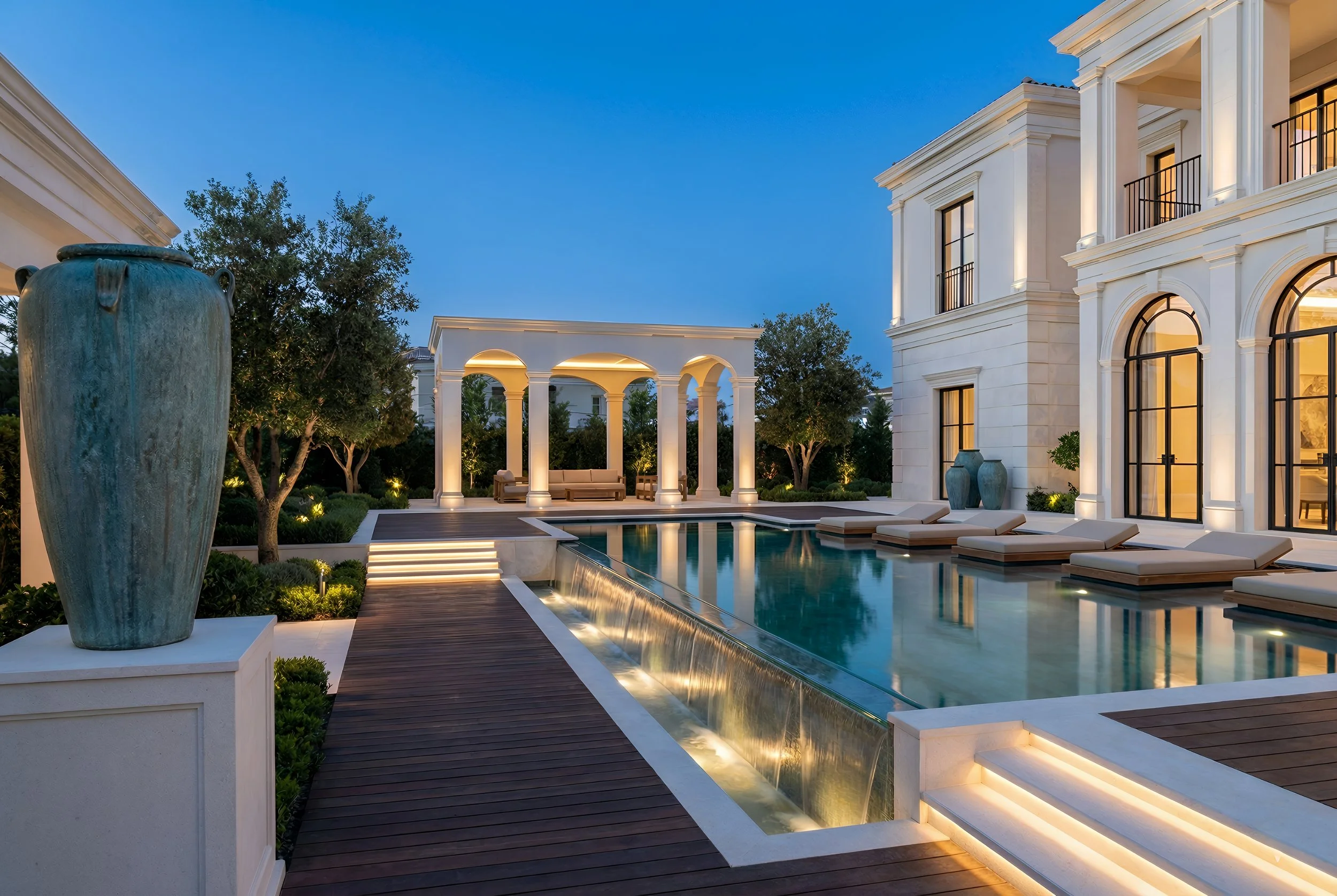 Luxury modern house with a backyard pool, lounge chairs, decorative vases, trees, and an illuminated pergola during twilight.