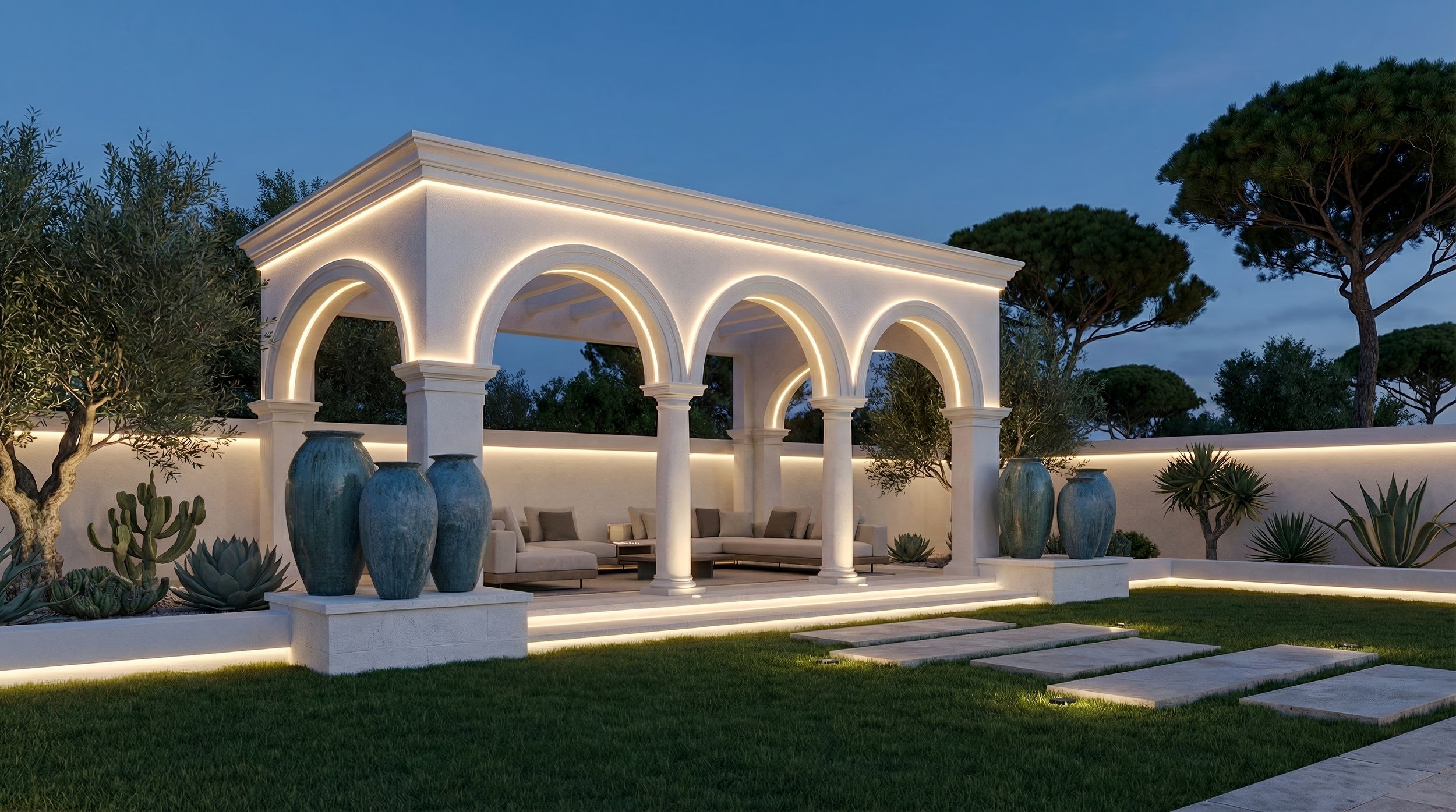 Nighttime view of an elegant outdoor patio with white arches and columns, illuminated with soft lighting. The patio has comfortable seating and large decorative vases, surrounded by lush greenery and trees.