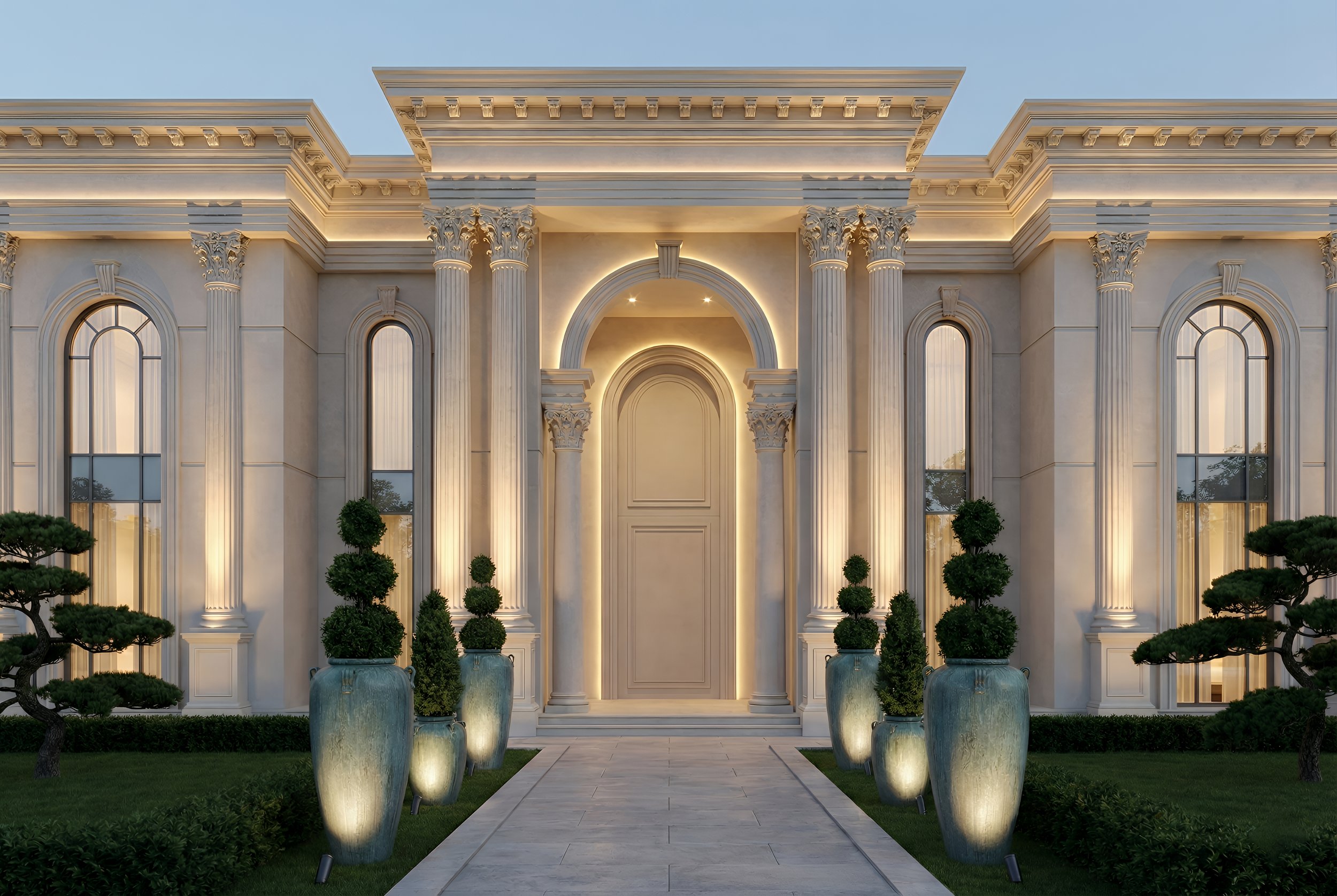 Front view of a grand, neoclassical-style mansion with columns, arched windows, and decorative arches, illuminated at night. Potted topiary trees line a stone pathway leading to the entrance.