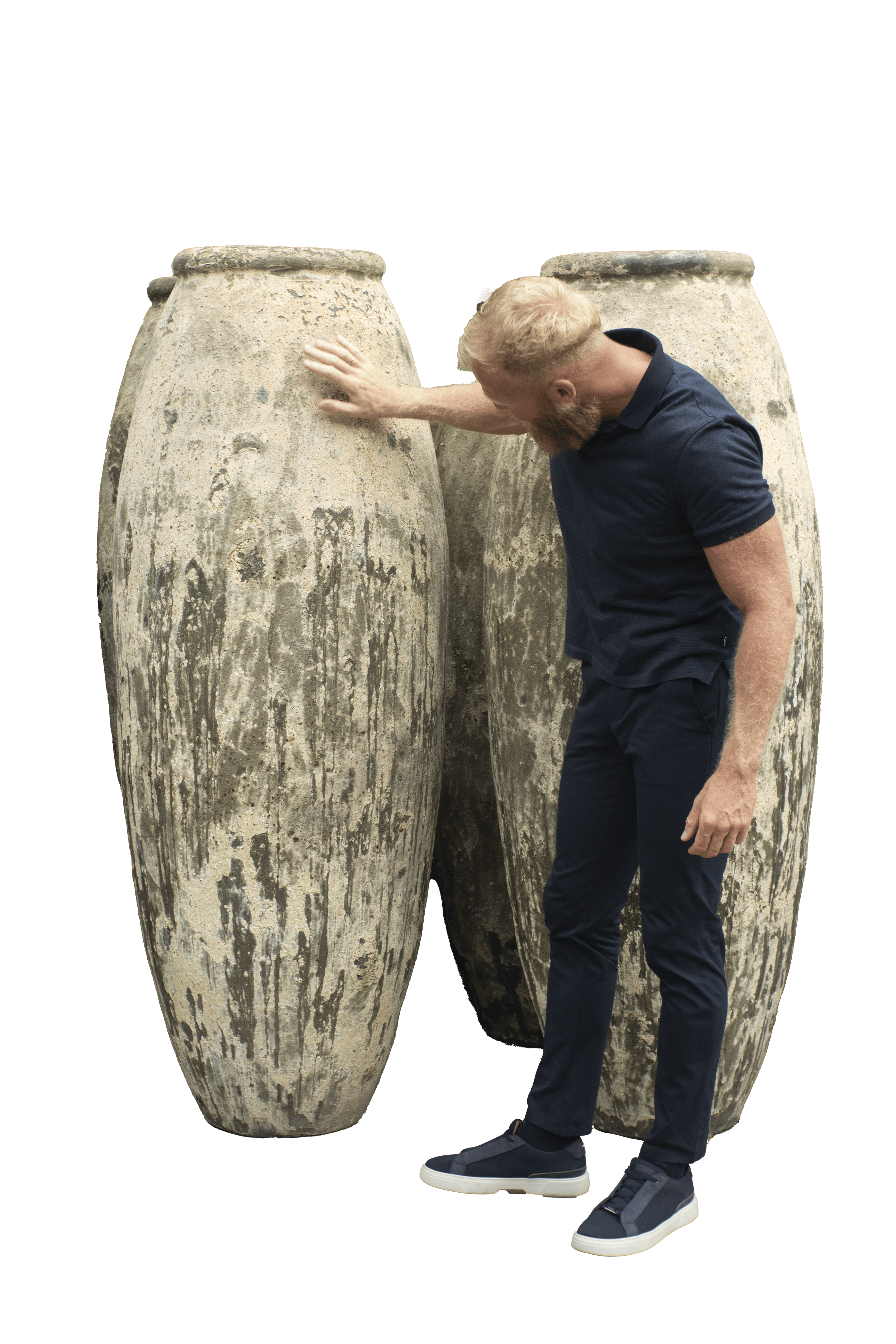 A man in dark clothing touching the surface of large, weathered stone sculptures that resemble elongated jars or vases.
