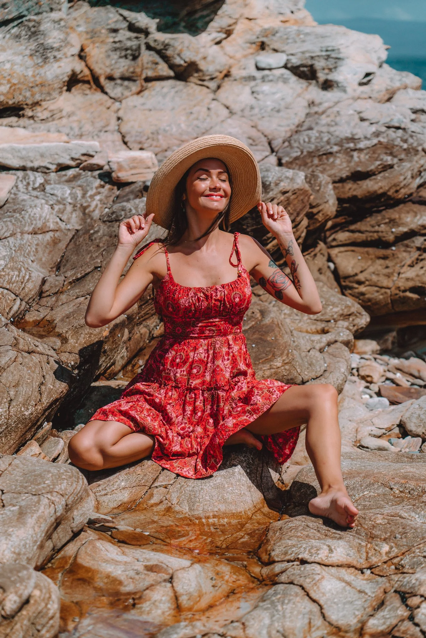 A woman in a red patterned dress and a wide-brimmed straw hat sitting on rocks near water, smiling with eyes closed.