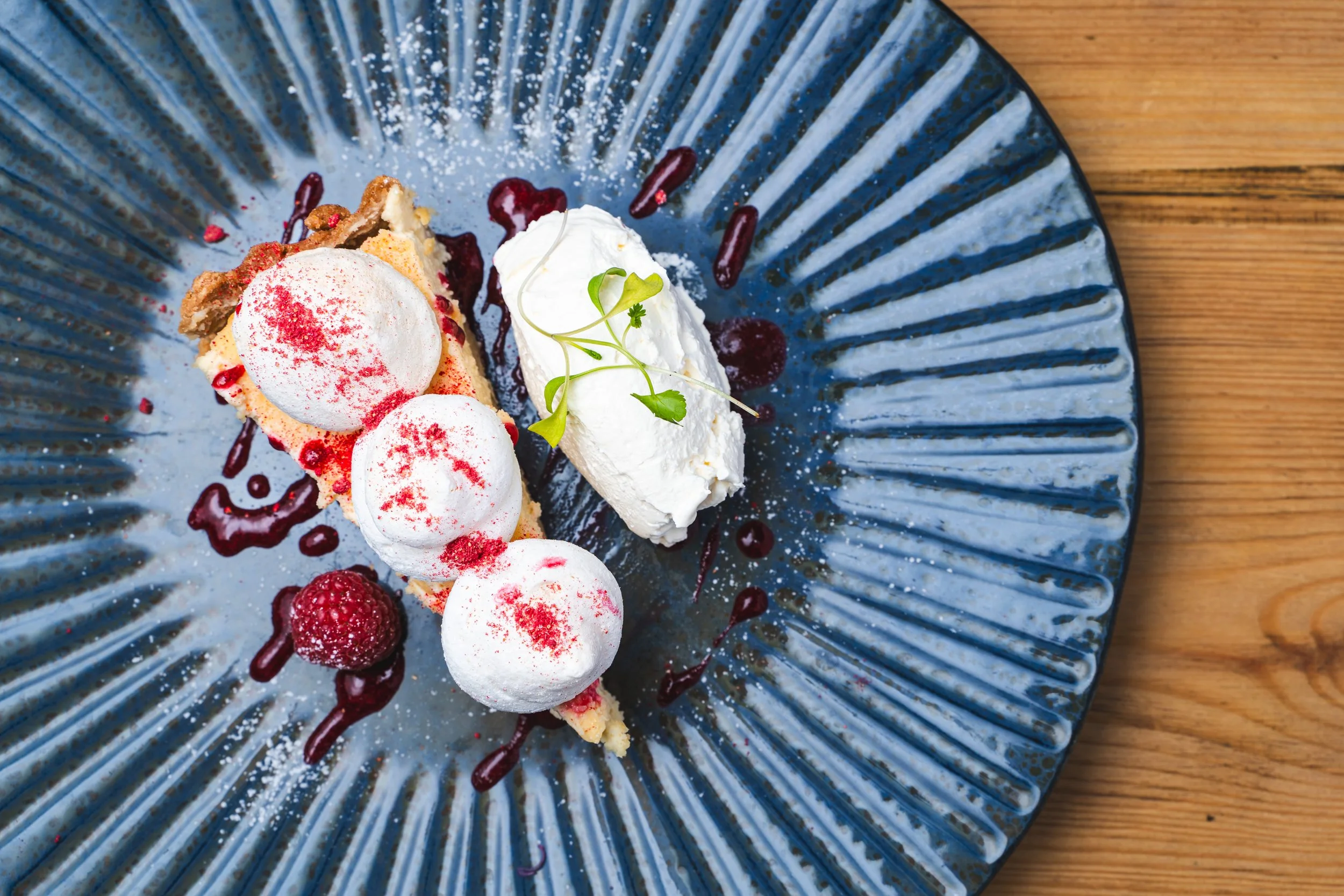 Dessert plate with a slice of tart topped with three meringue dollops and a raspberry, garnished with a sprig of microgreens, drizzled with berry sauce, served on a blue textured plate on a wooden surface.