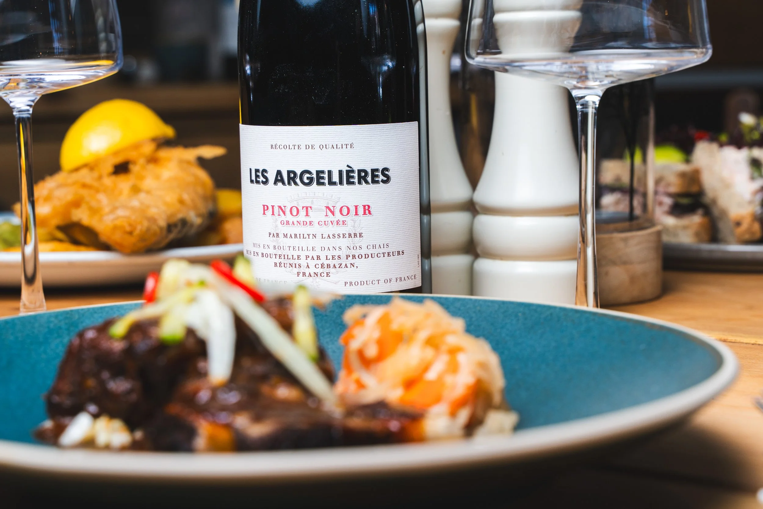 A bottle of Les Argelières Pinot Noir wine in the center with two wine glasses nearby. In the background, there's a plate with fried fish garnished with lemon and some side dishes. There is also a dish with mashed sweet potatoes topped with sliced green onions.