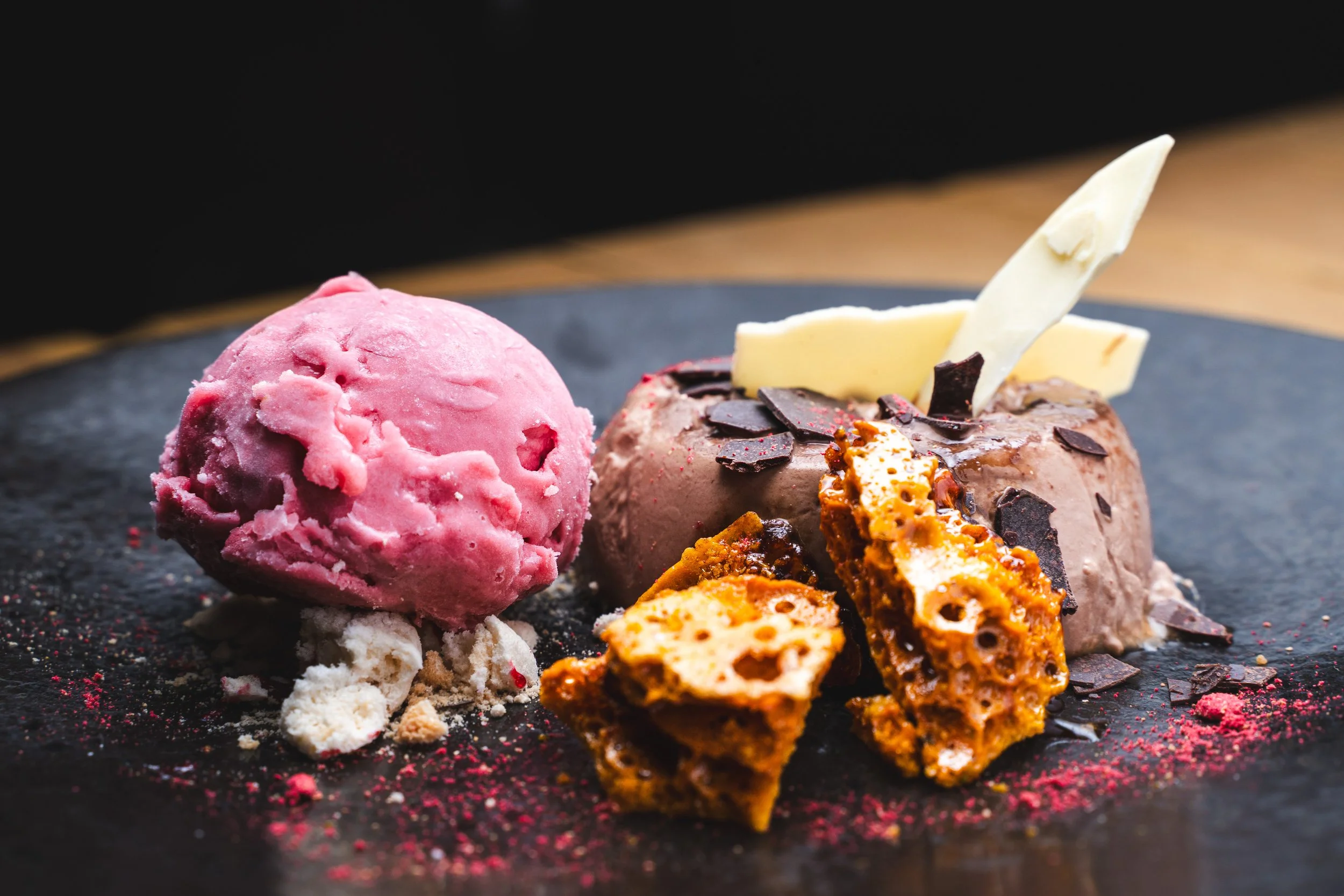 A dessert plate with a scoop of pink ice cream, a chocolate dessert garnished with white chocolate and chocolate shavings, and caramel brittle pieces, on a dark surface.