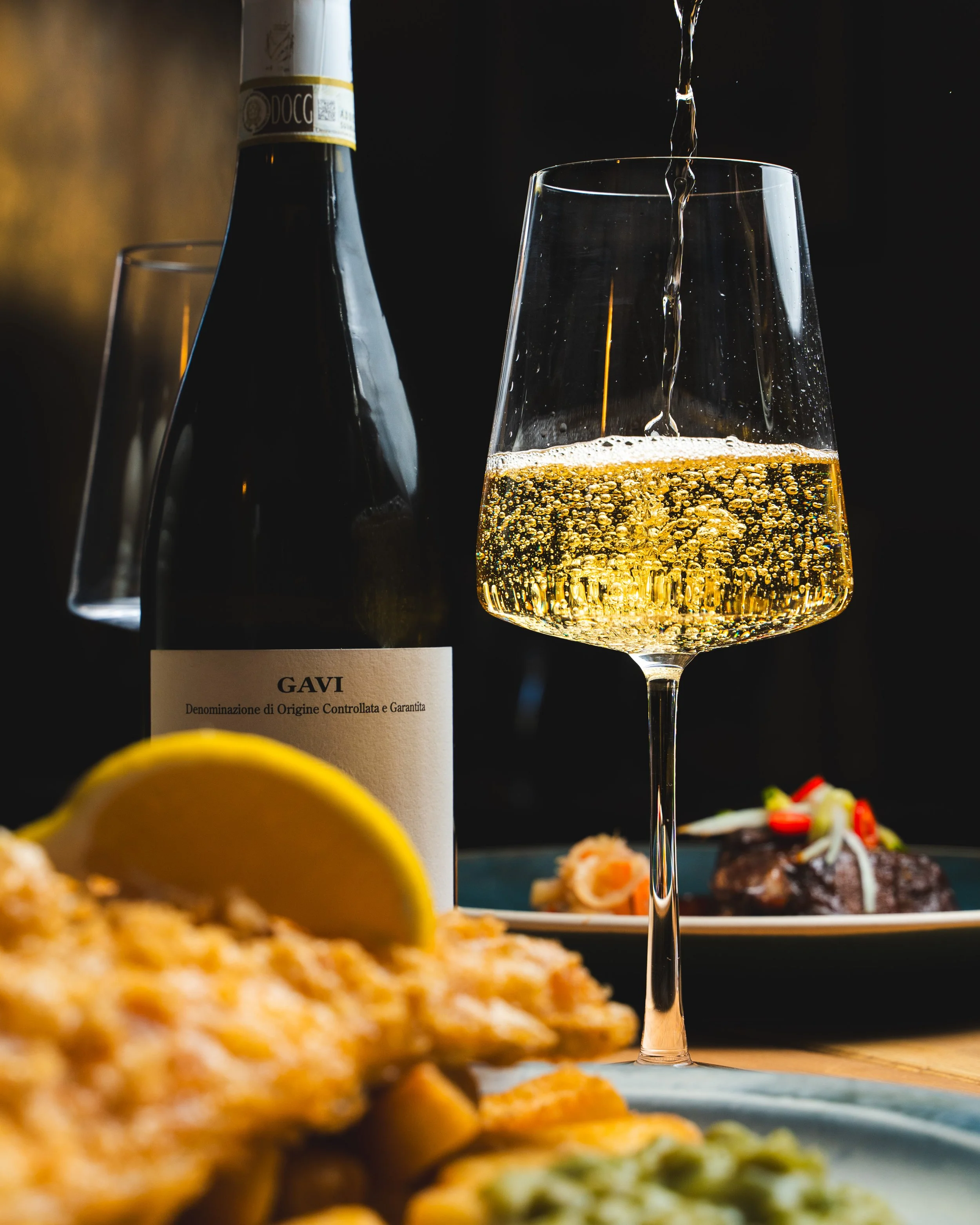 A glass of sparkling white wine being poured, a dark bottle of Gavi wine, a lemon wedge, fried food, and a plate with a piece of meat with garnishes in the background.