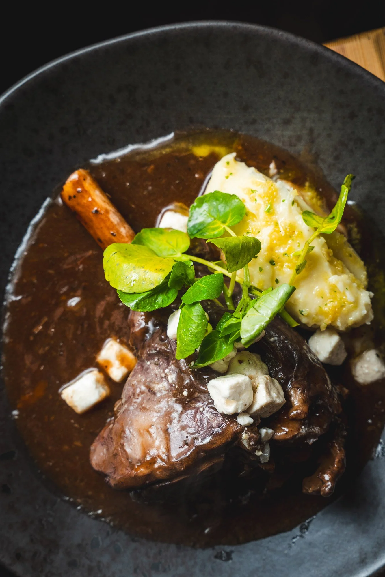 Lamb stew with mashed potatoes, a sprig of herbs, and a cinnamon stick in a black bowl.