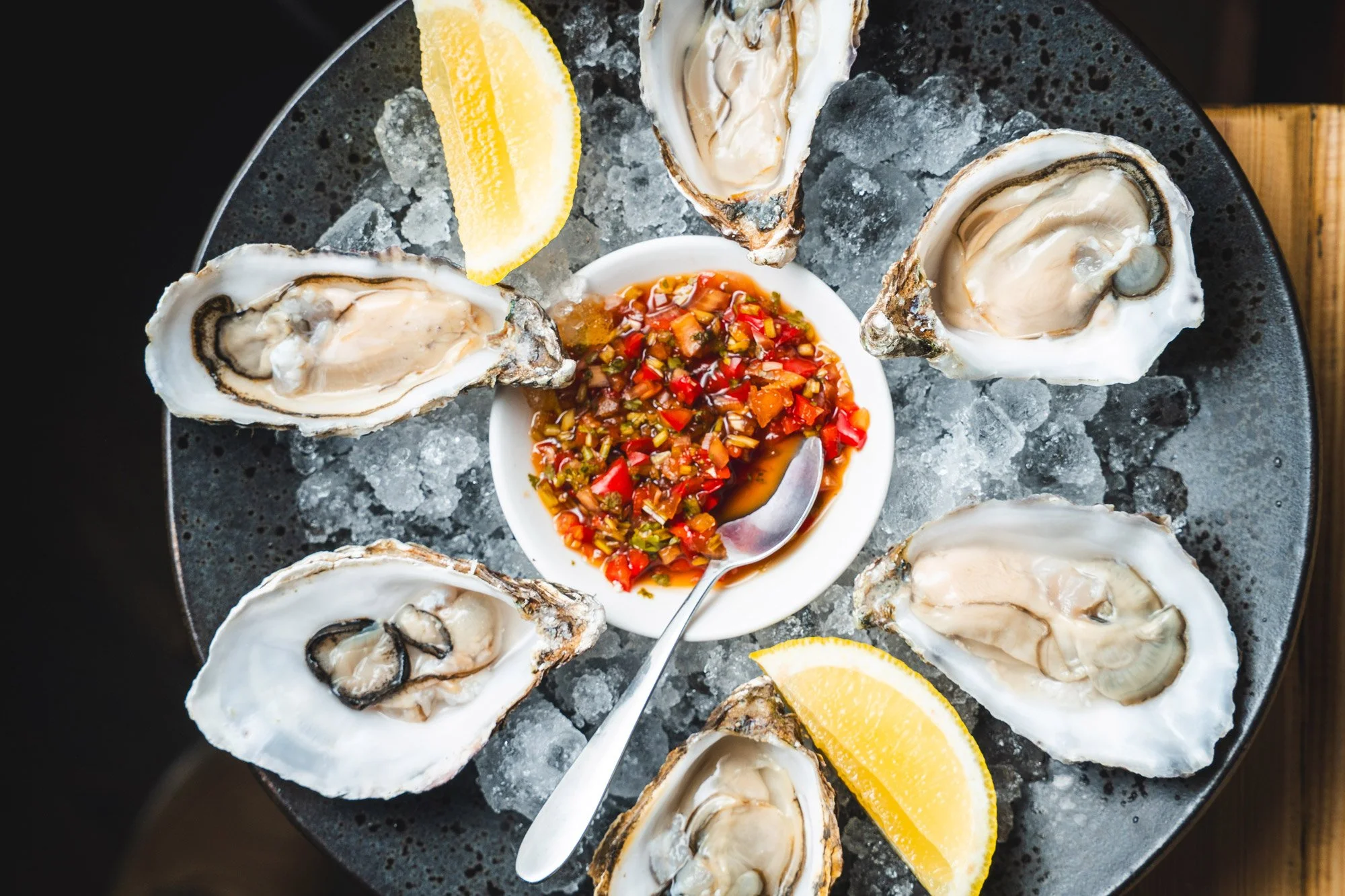 Six opened oysters on ice arranged in a circle on a dark tray, with lemon wedges and a small bowl of spicy red cocktail sauce in the center.