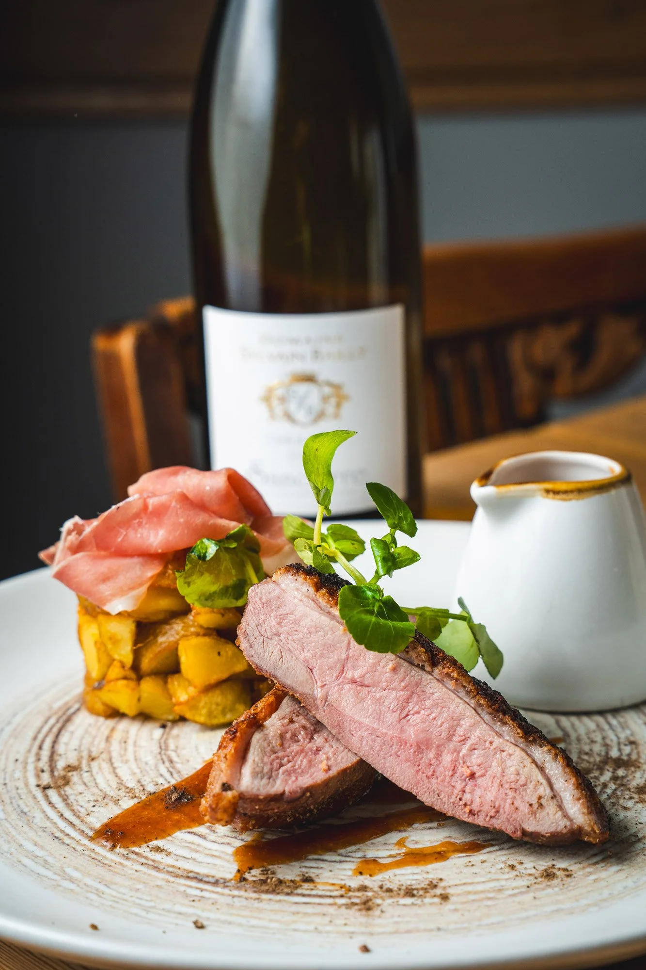 A gourmet plate with sliced, cooked meat, garnished with greens, served with roasted potatoes topped with prosciutto, and a small creamer pitcher, with a bottle of wine in the background.