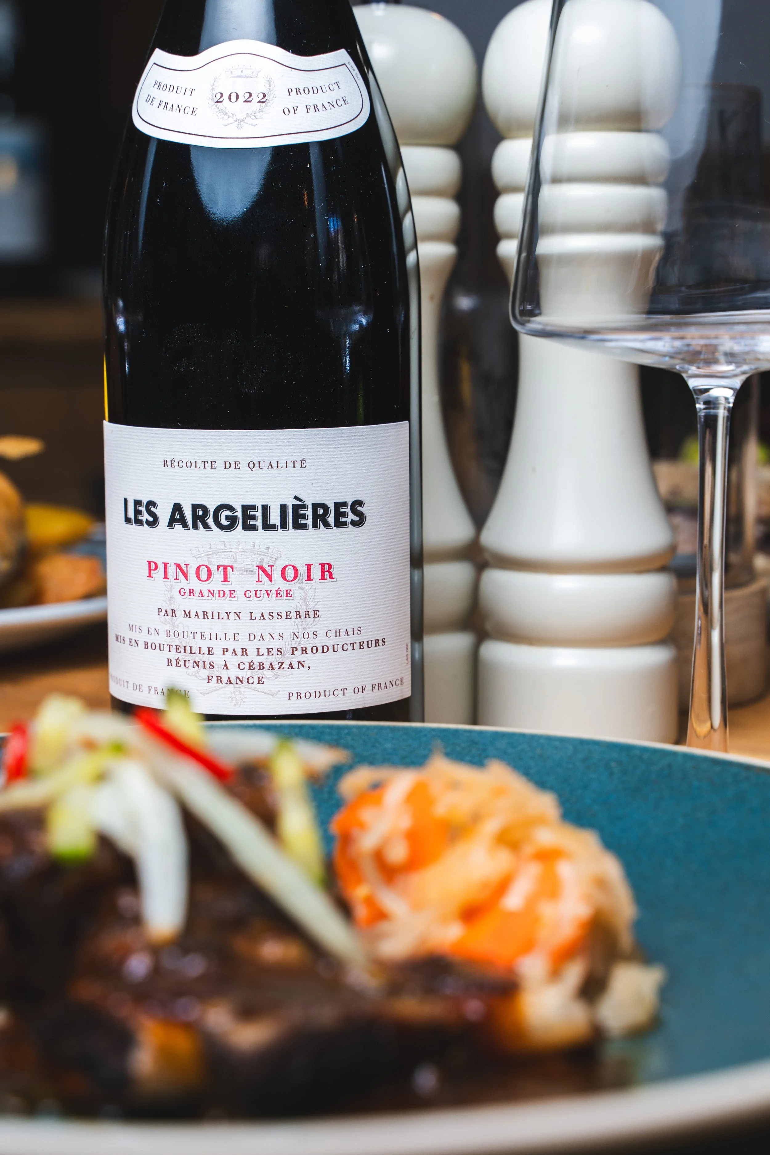 A bottle of Les Argelieres Pinot Noir wine with a glass beside it, on a table with blurred food in the foreground.