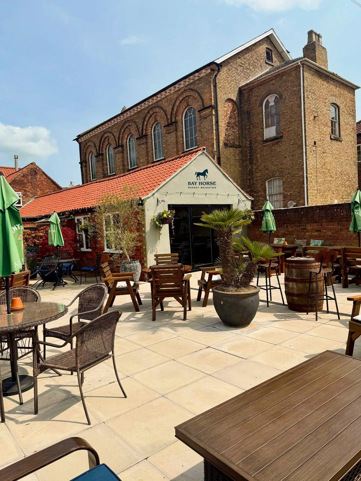 Coffee shop patio with tables, chairs, umbrellas, and potted plants, with a brick building in the background and a sign that reads 'Bay Horse Market Weighton'.