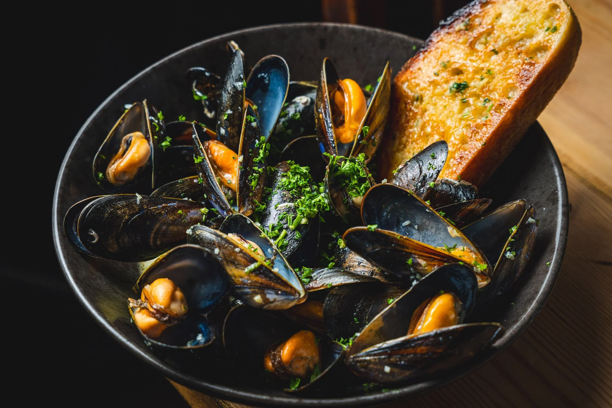 A black bowl filled with cooked mussels garnished with chopped herbs, accompanied by a slice of toasted garlic bread on the side.
