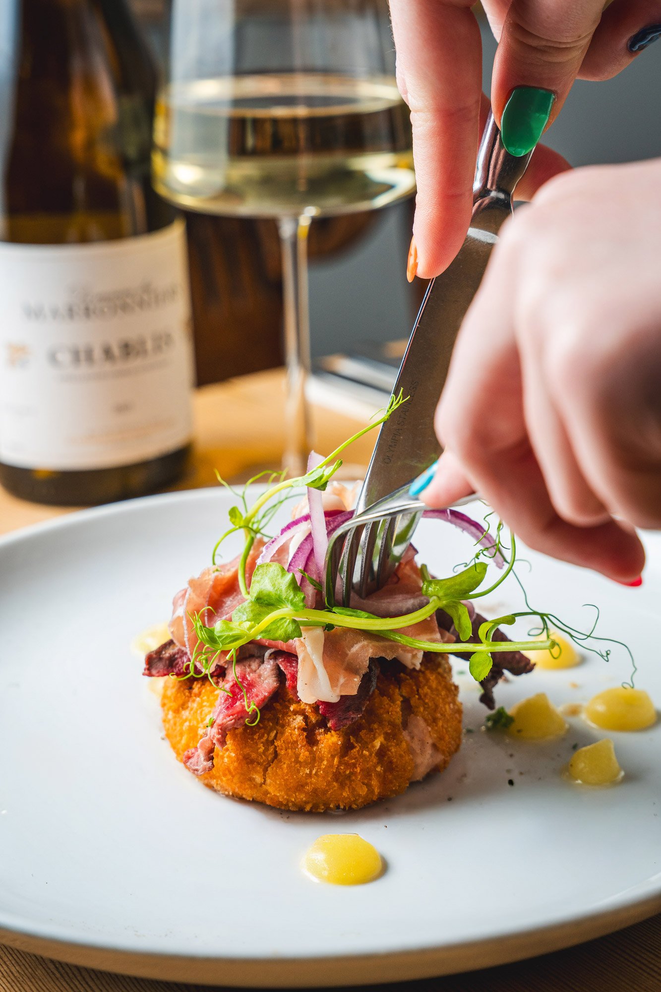 Hand cutting into a plated gourmet dish with microgreens and sauce droplets, with a glass of white wine and a wine bottle in the background.
