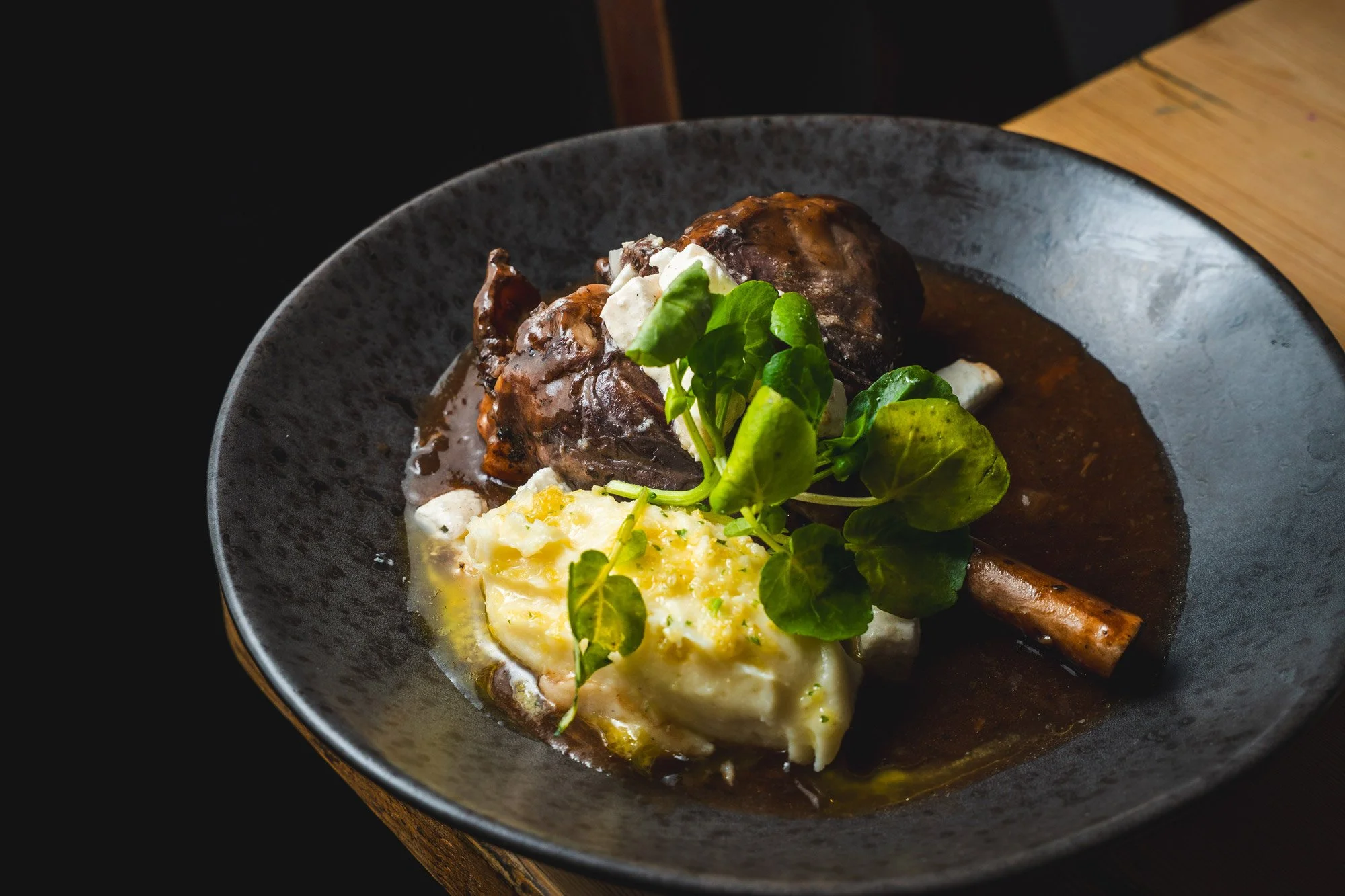 A plated dish featuring a succulent braised beef shank with sauce, served with mashed potatoes and garnished with fresh greens.