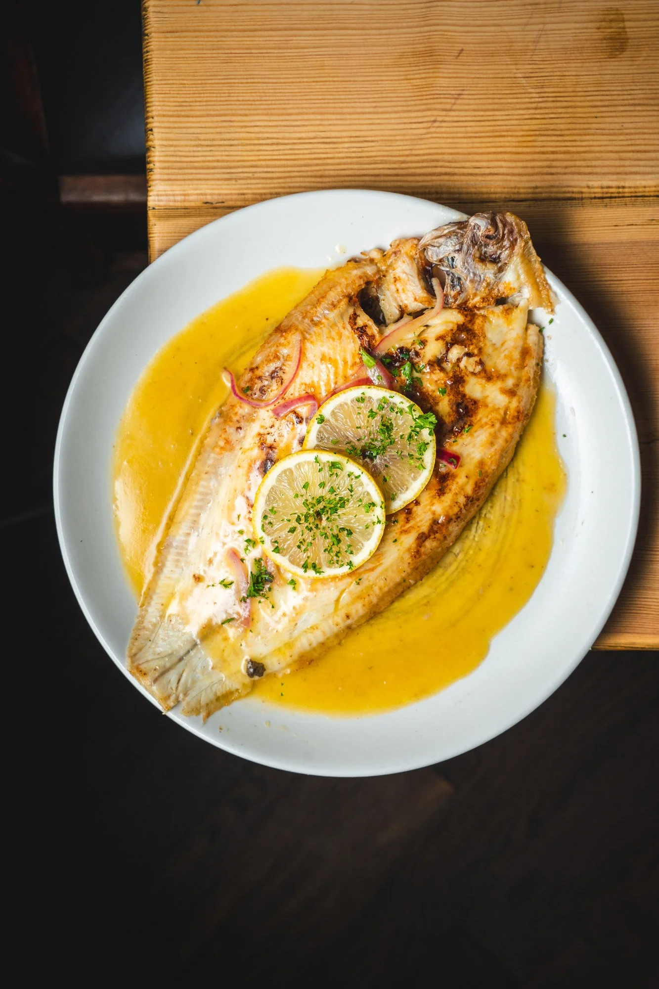 Grilled fish with lemon slices, garnished with parsley, served with a yellow sauce on a white plate on a wooden table.