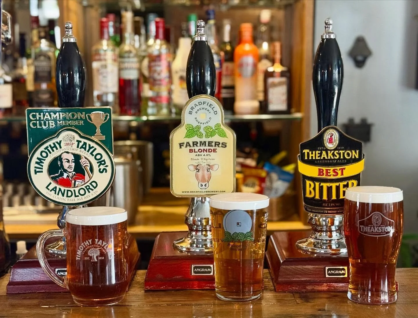Three beer taps with labeled handles and three glasses of beer on a wooden bar, with a blurred background of liquor bottles.