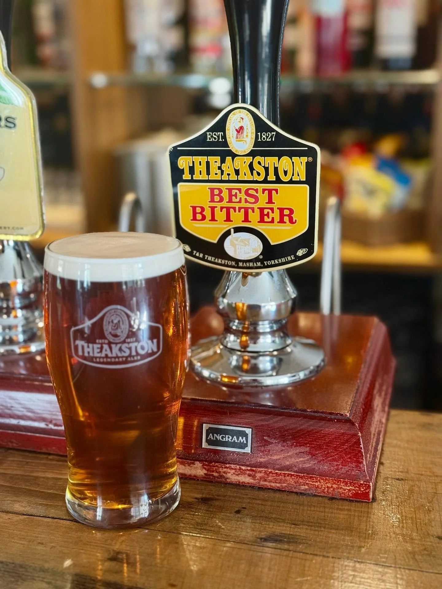 A glass of beer with a frothy head sitting on a wooden table in front of a bar tap that advertises Theakston Best Bitter, with a logo and name on the glass and tap.