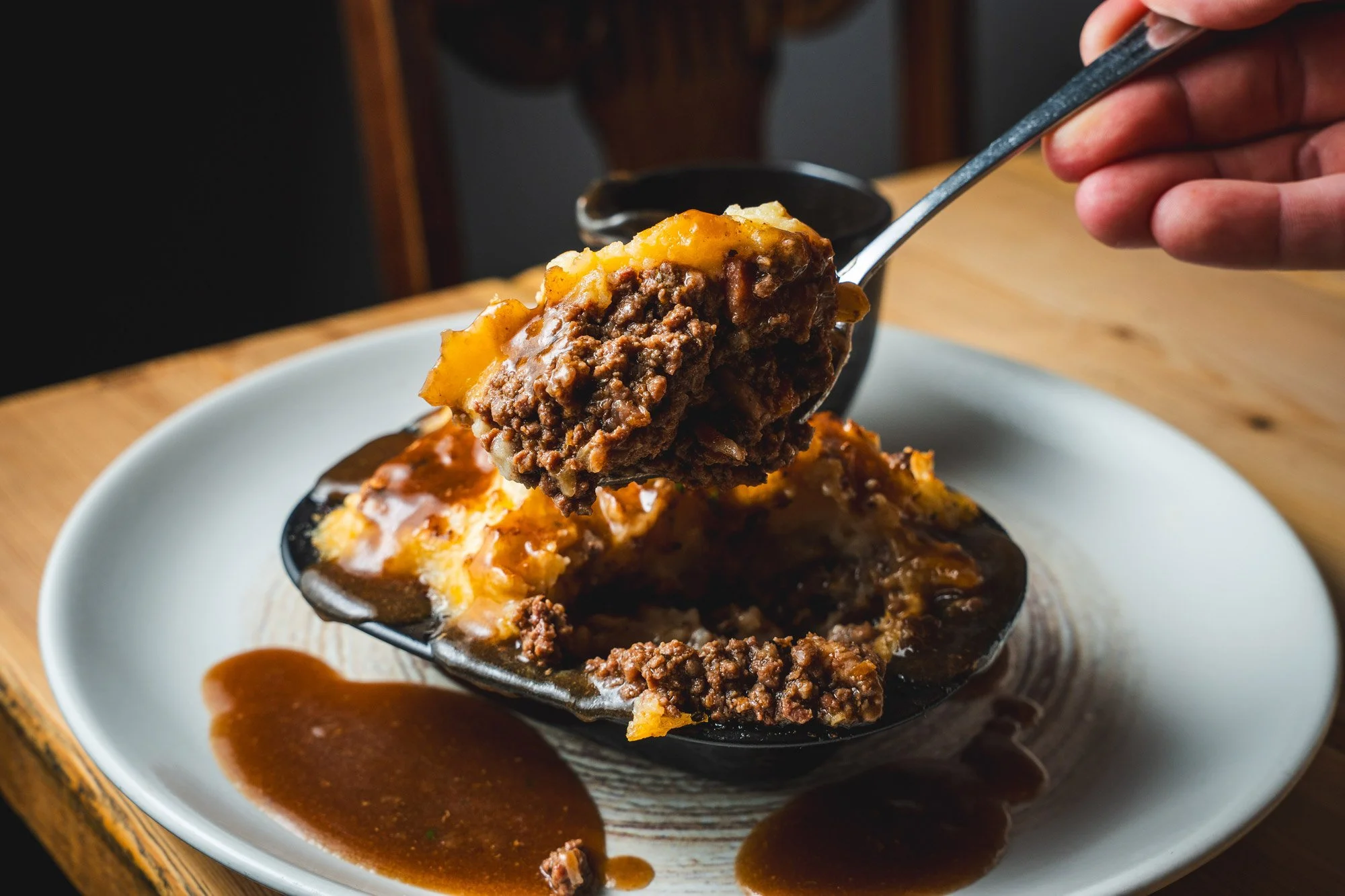 A spoonful of shepherd's pie being lifted from a black bowl with cooked ground beef, mashed potatoes, and gravy, on a white plate with additional gravy around the bowl.