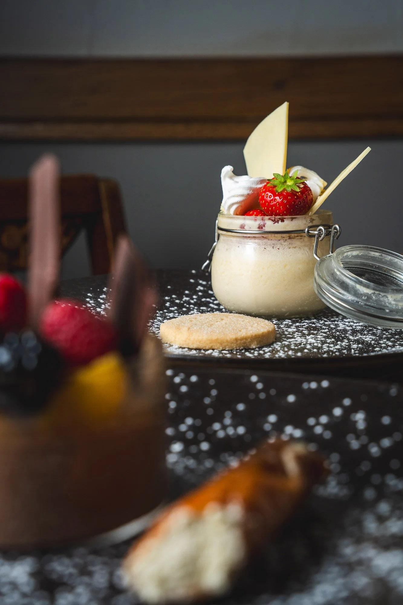 Glass jar with strawberries, whipped cream, and white chocolate shards, sitting on a black surface dusted with powdered sugar, with a cookie nearby.