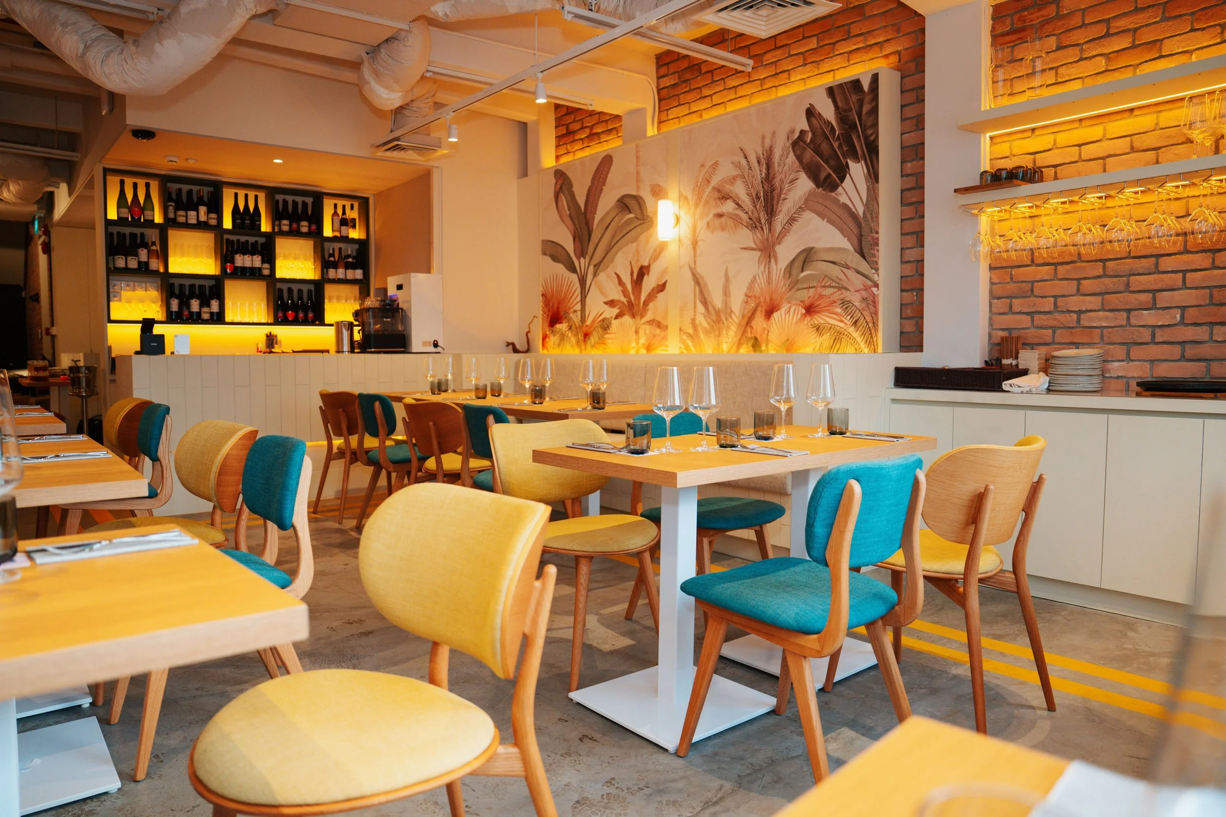 Interior of a modern restaurant with colorful chairs, wine glasses, a brick wall, and a large tropical mural.