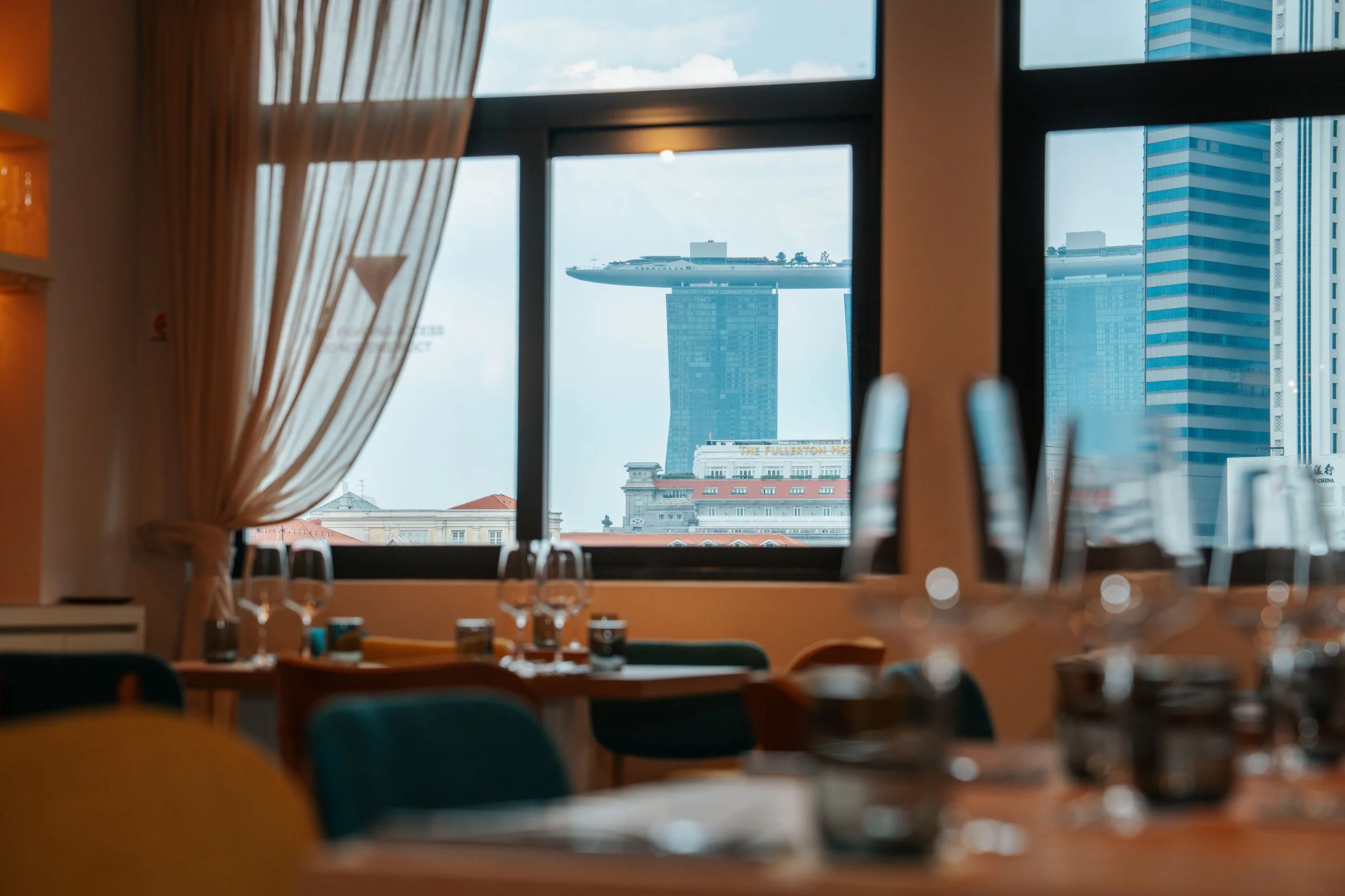 Indoor dining area with tables set with glasses and utensils, large window showing the Marina Bay Sands hotel and city skyline in Singapore.