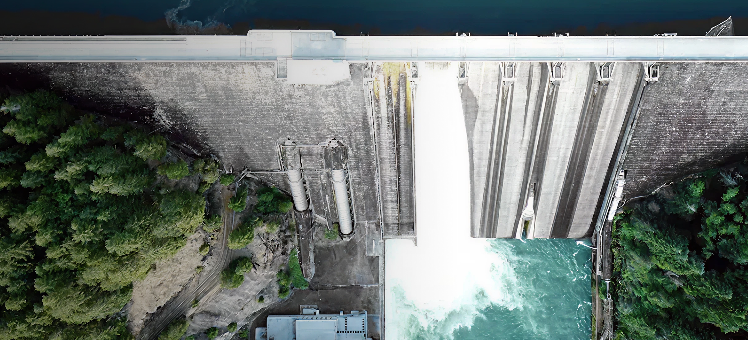 An aerial view of a dam releasing water with water flowing down its face and into the river below, surrounded by green trees.