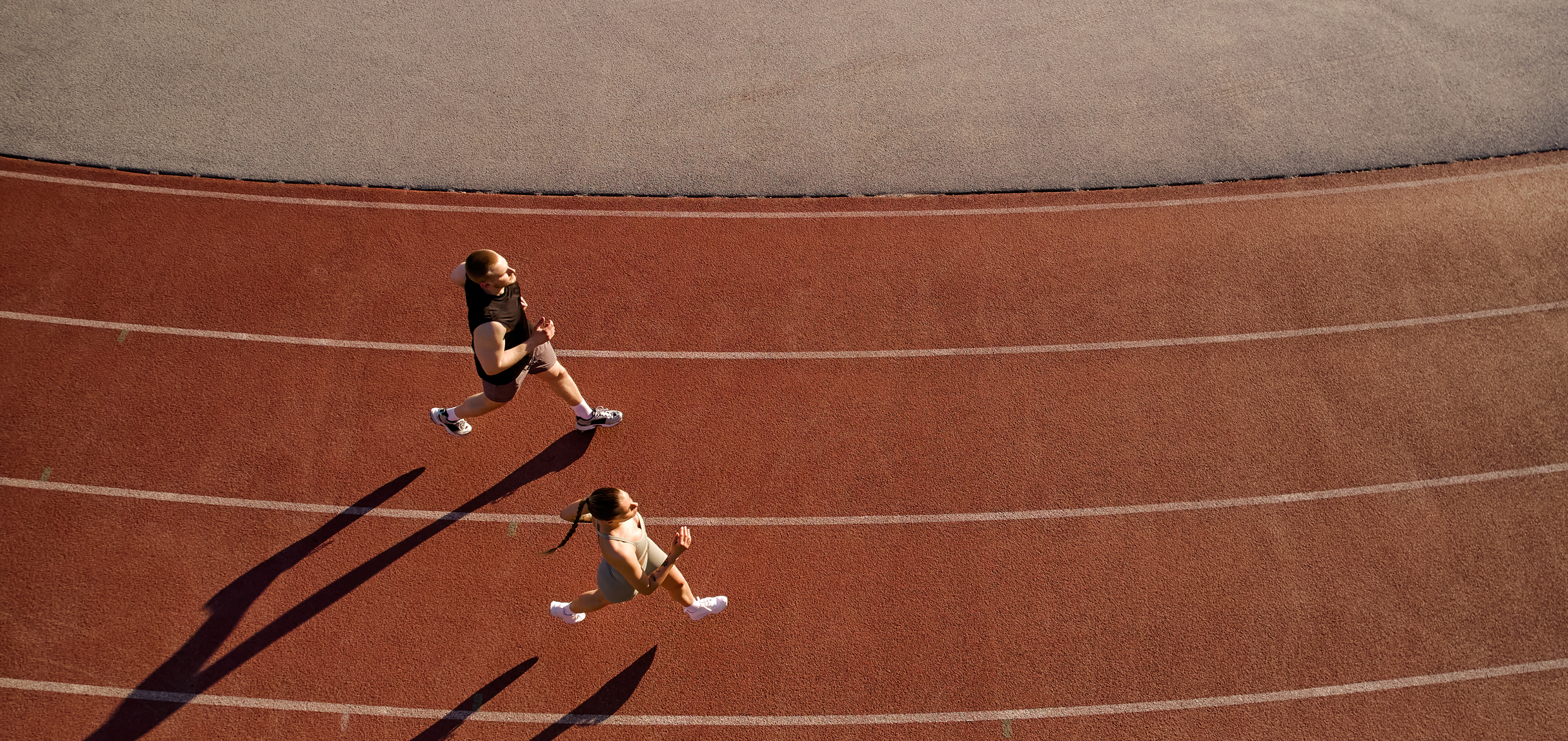 Two runners jog on a red outdoor track from an overhead view, casting long shadows.