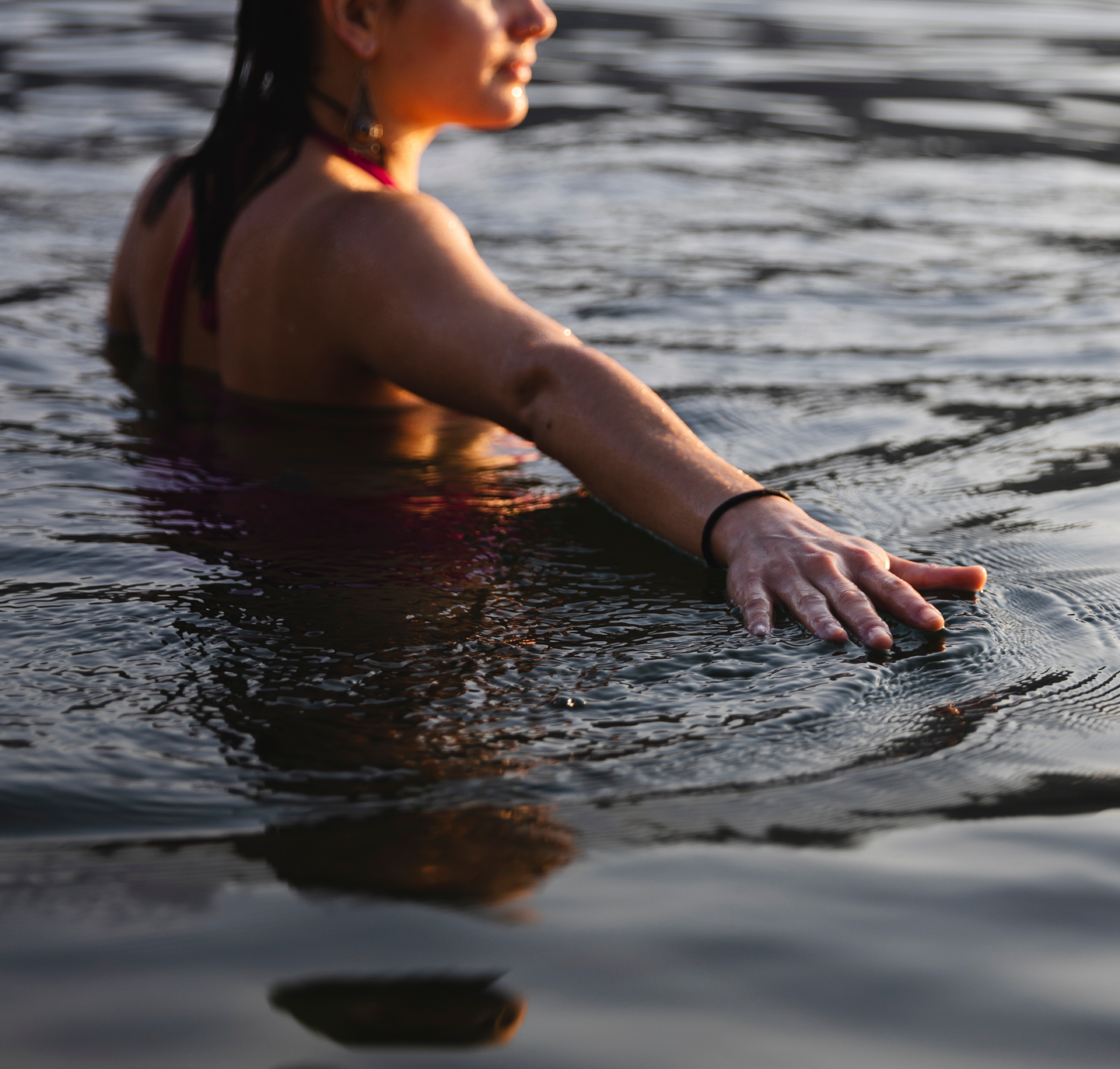 A woman in a red swimsuit partially submerged in water, reaching out with her right arm, during sunset or sunrise.