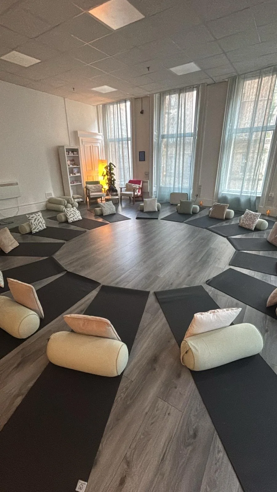 A spacious room set up for yoga or meditation with black mats, bolsters, and pillows arranged in a circle, large windows with sheer curtains allowing natural light, and cozy chairs and shelves in the background.