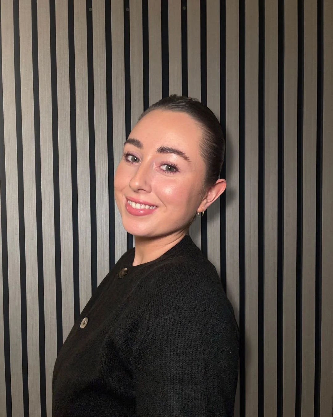 Portrait of a woman with dark hair pulled back, smiling, wearing a black top, standing against a black and gray striped wall.