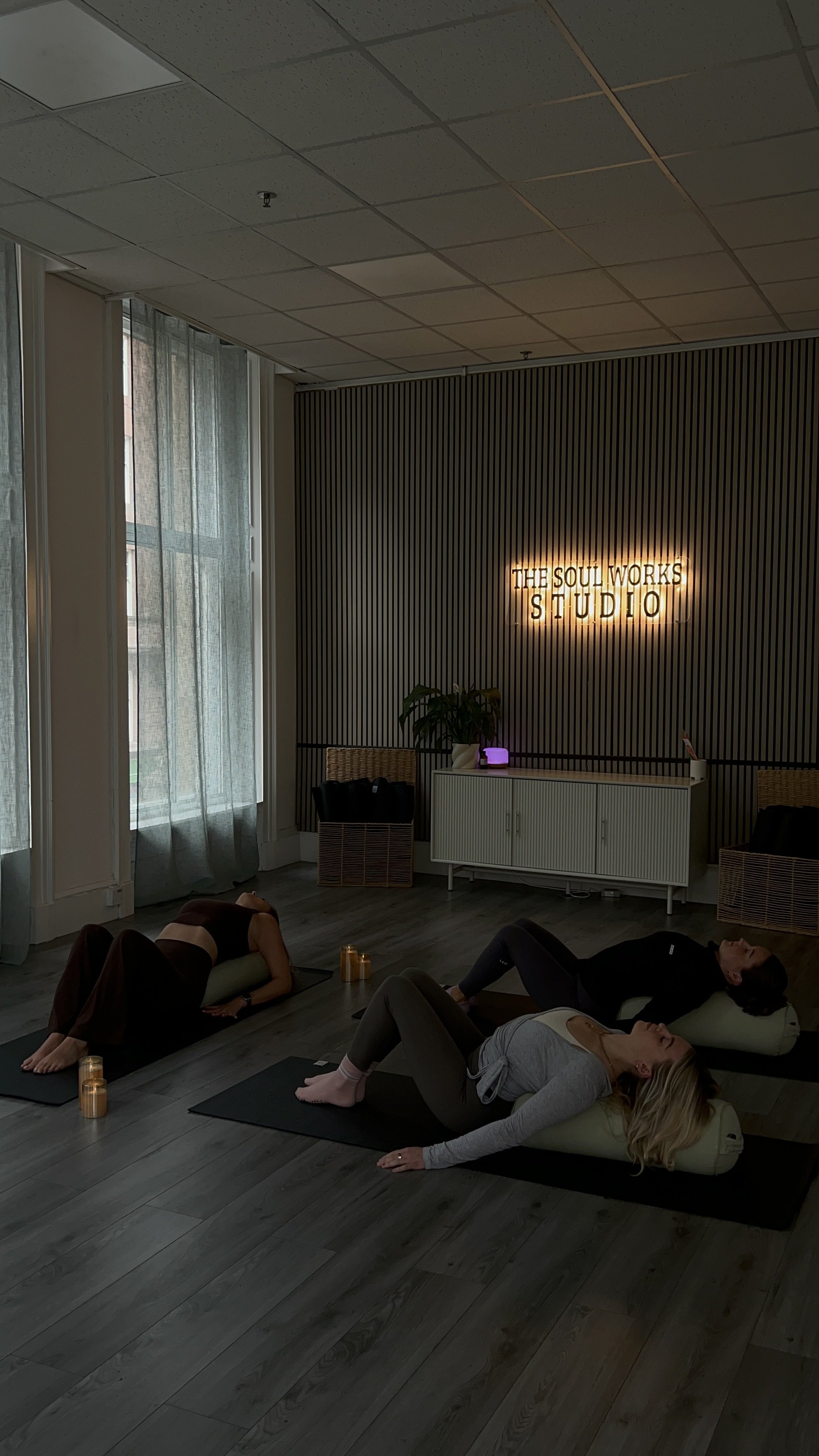 Three women practicing yoga in a studio along a wall with a neon sign that reads 'The Soul Works Studio'.