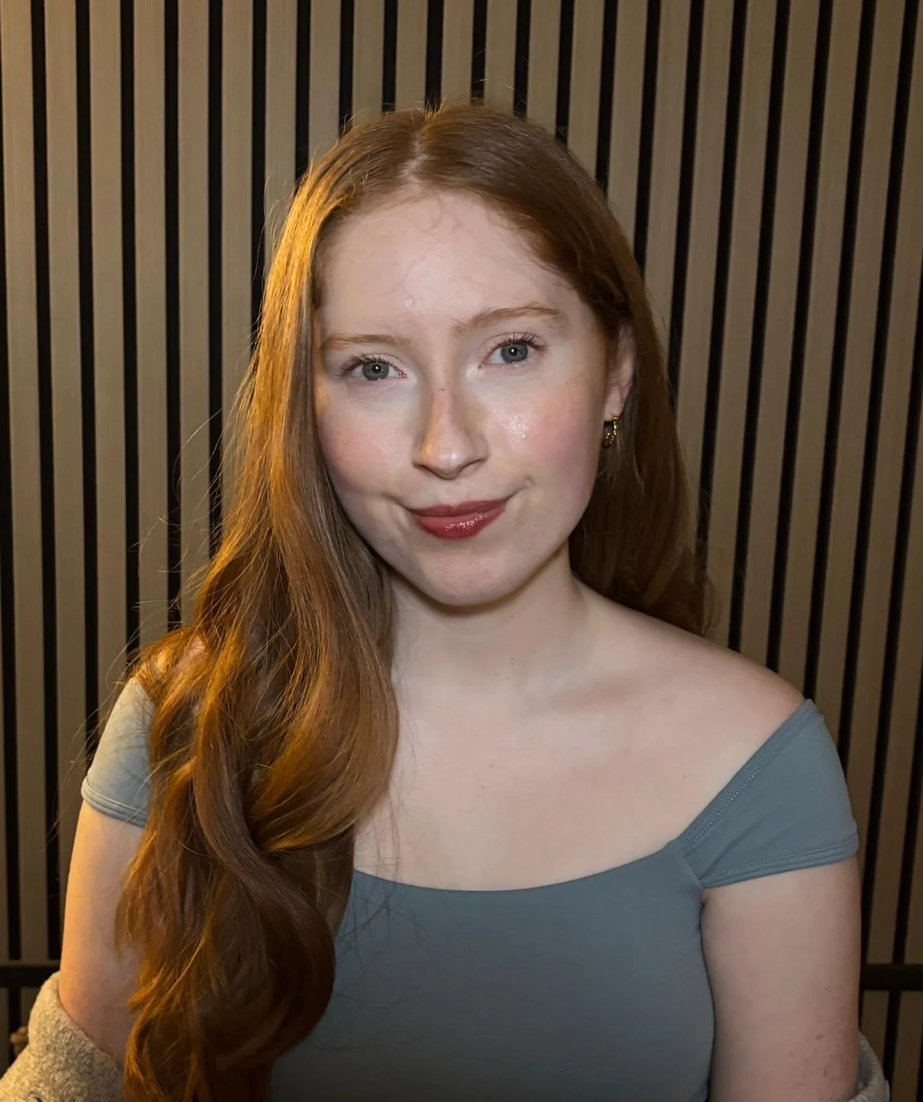 A young woman with red hair, blue eyes, and fair skin smiling at the camera, wearing a light blue top and small earrings, against a background of vertical black wooden slats.