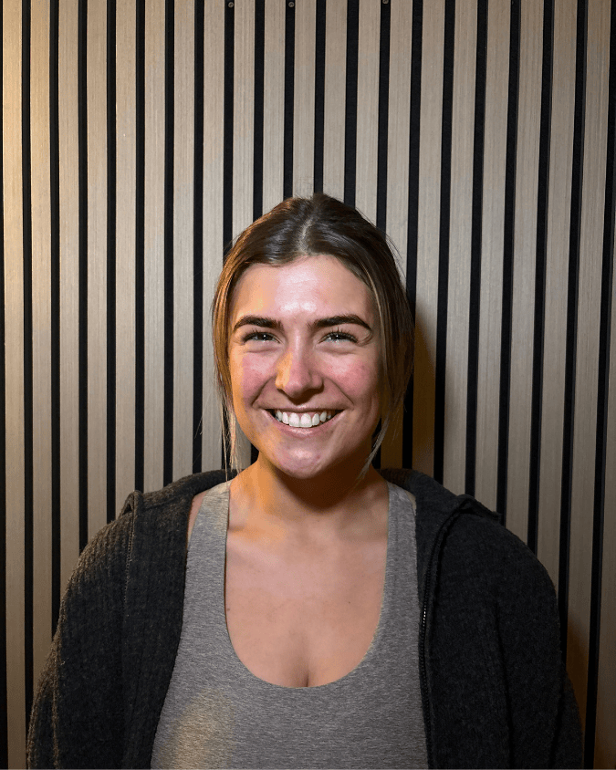 Smiling woman with dark hair pulled back, wearing a gray top and black zip-up hoodie, standing in front of a black and beige striped wall.