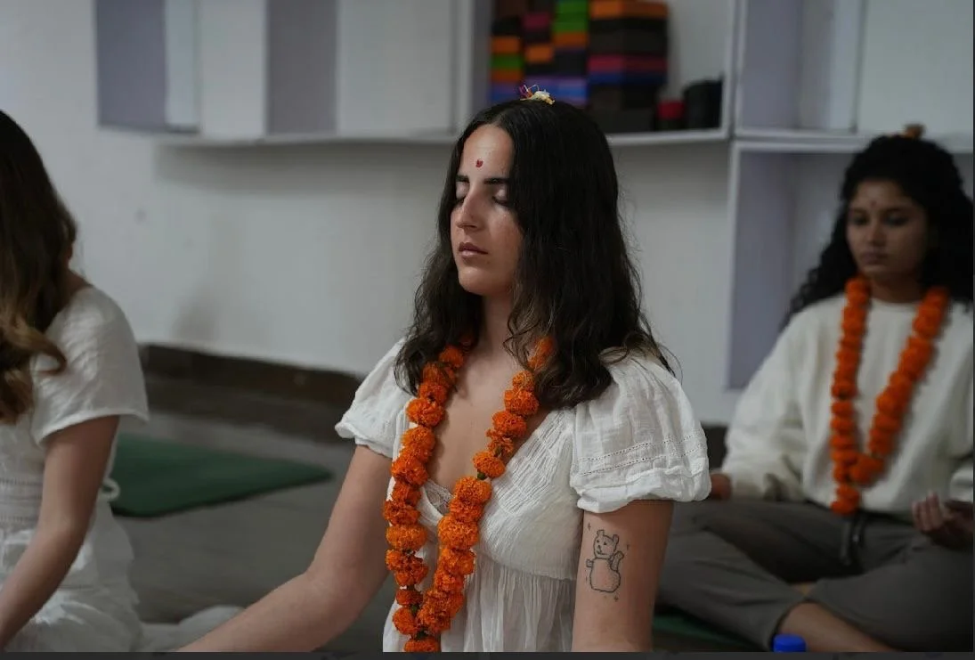 A woman with dark hair practicing meditation, wearing a white dress and an orange marigold garland, with her eyes closed and an elephant tattoo on her arm, in a room with others also meditating.
