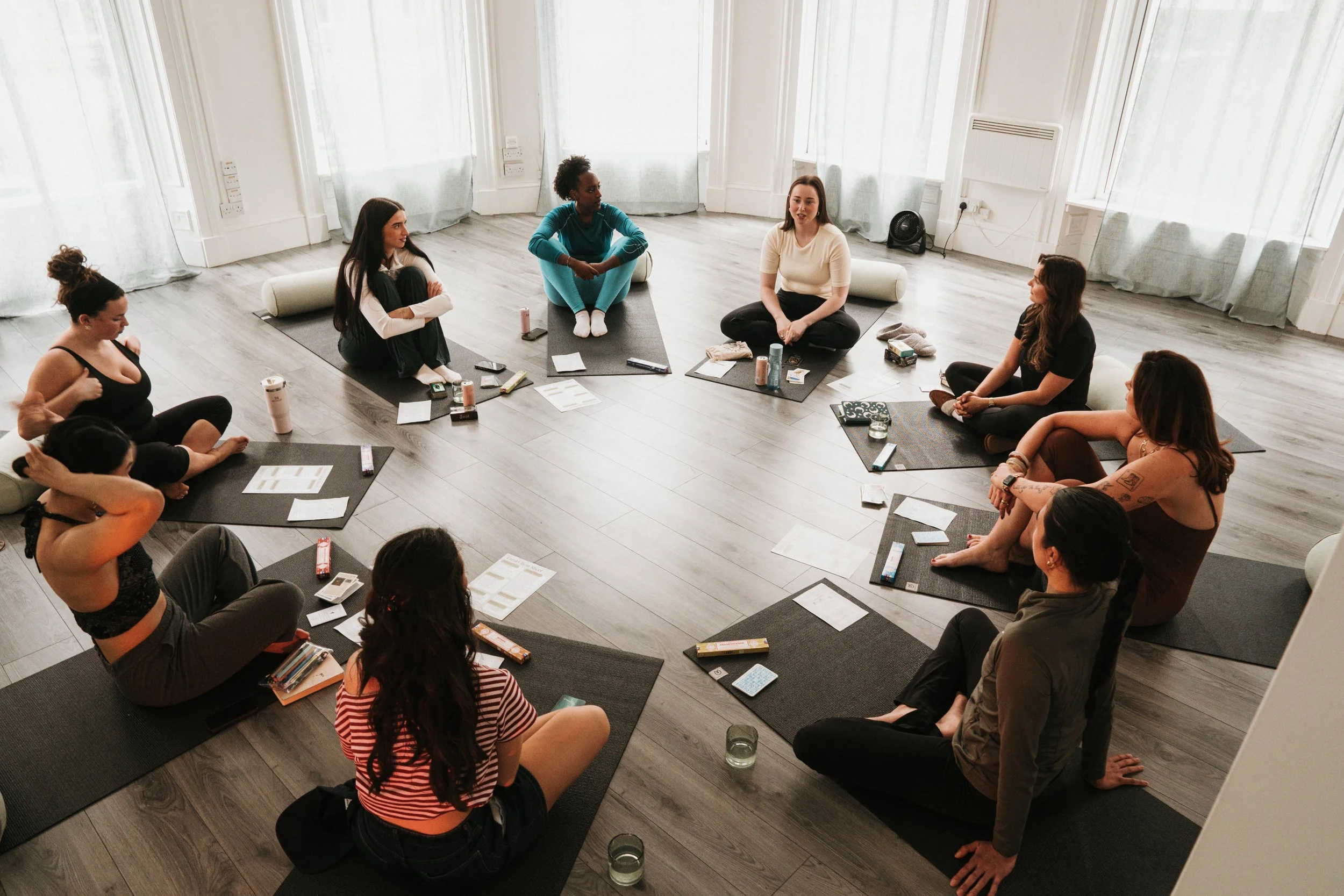 A group of women sitting cross-legged on yoga mats in a circle, engaging in a discussion or meditation session in a bright, spacious room.