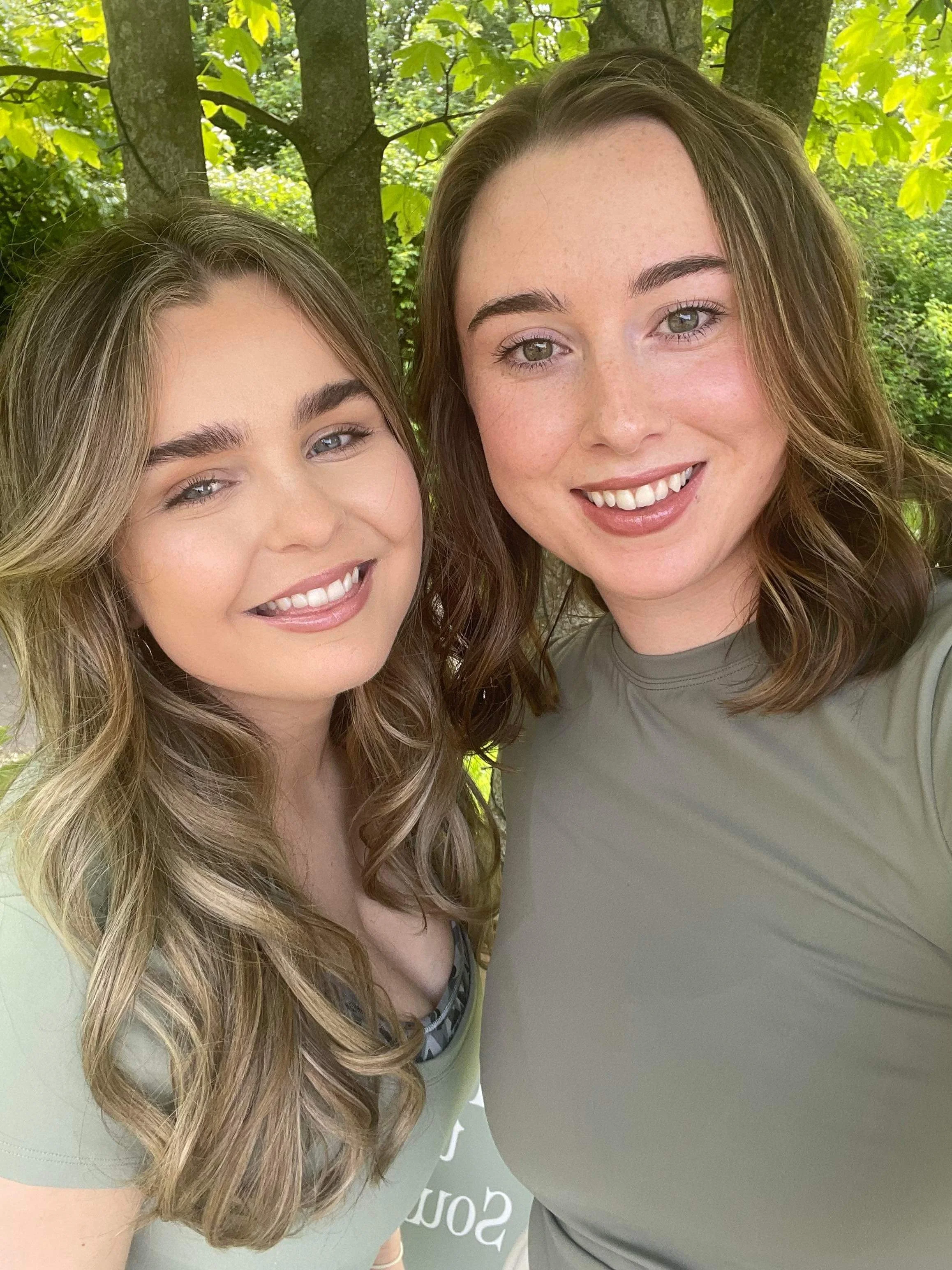 Two young women with long hair smiling outdoors in front of green trees.