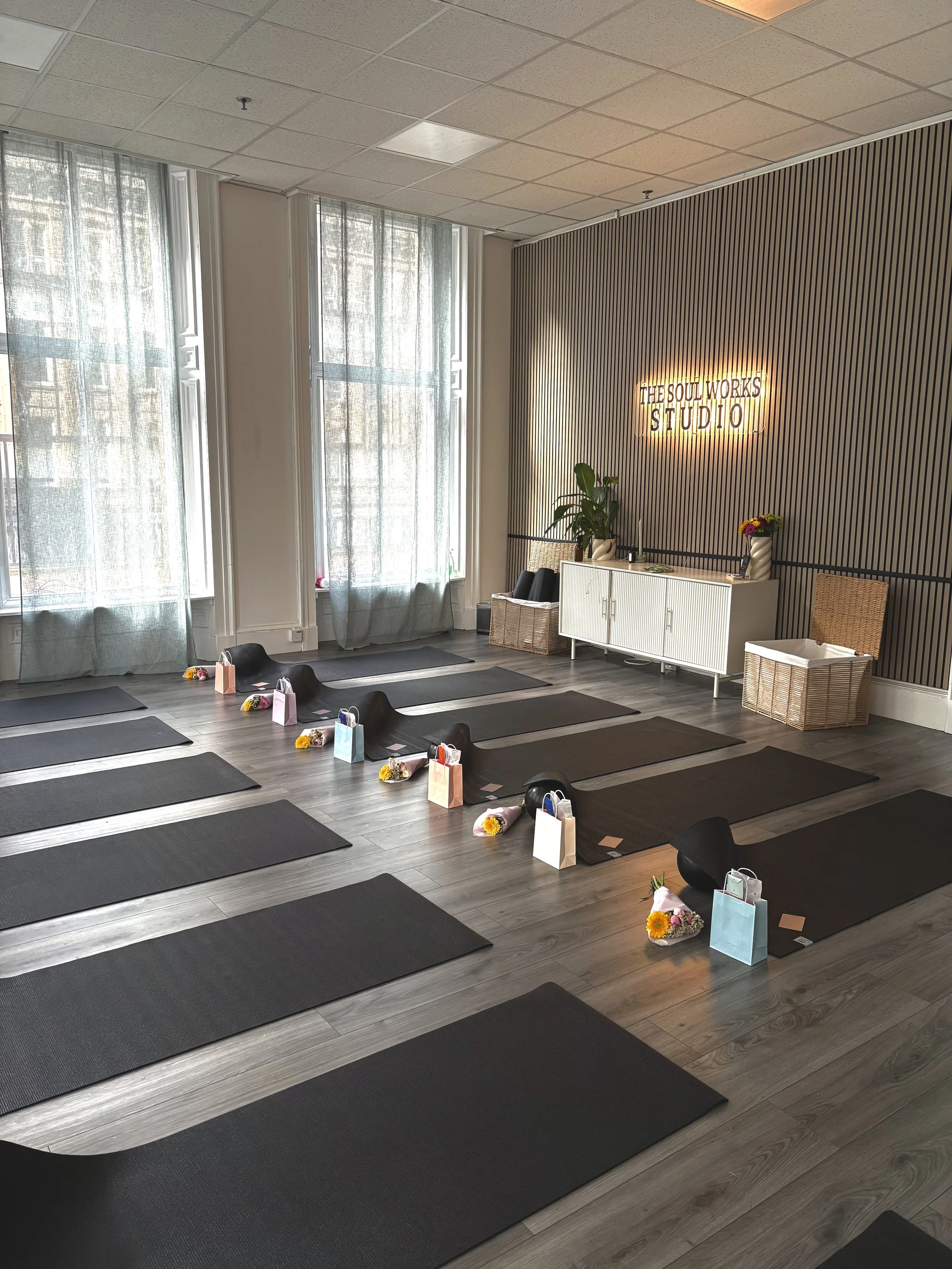 Yoga mats arranged in a grid on a wooden floor in a bright, airy studio with large windows, decorated with gift bags, flowers, and a neon sign that reads 'The Soul Works Studio' on a textured, striped wall.
