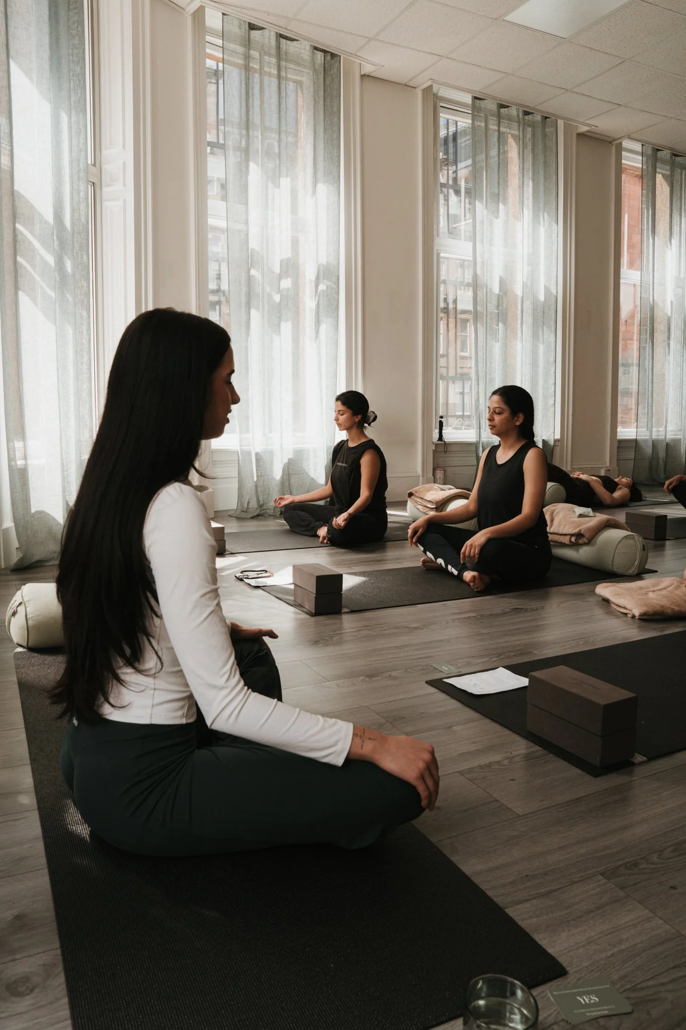 Group of women practicing meditation in a bright room with tall windows and sheer curtains, sitting cross-legged on yoga mats.