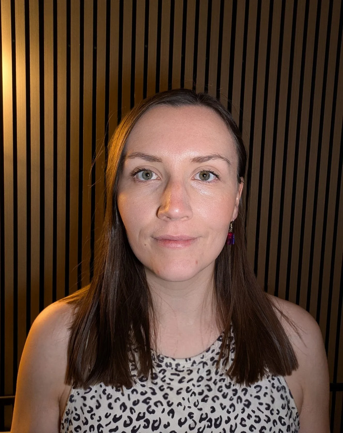 A woman with straight brown hair, wearing a sleeveless leopard print top and purple earrings, standing against a dark wooden slatted wall.